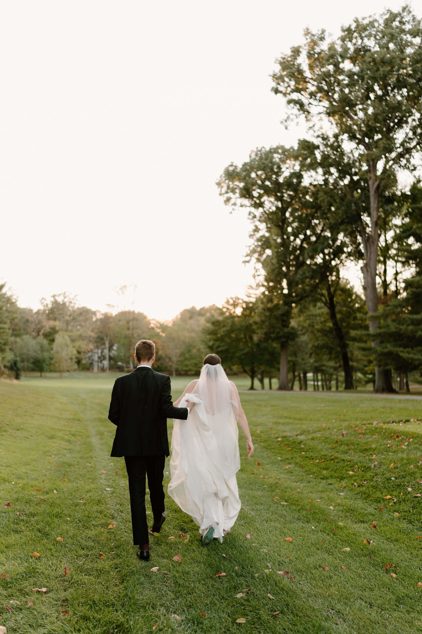 bride and groom walking along a grassy field, away from the camera, with the groom holding his bride's train