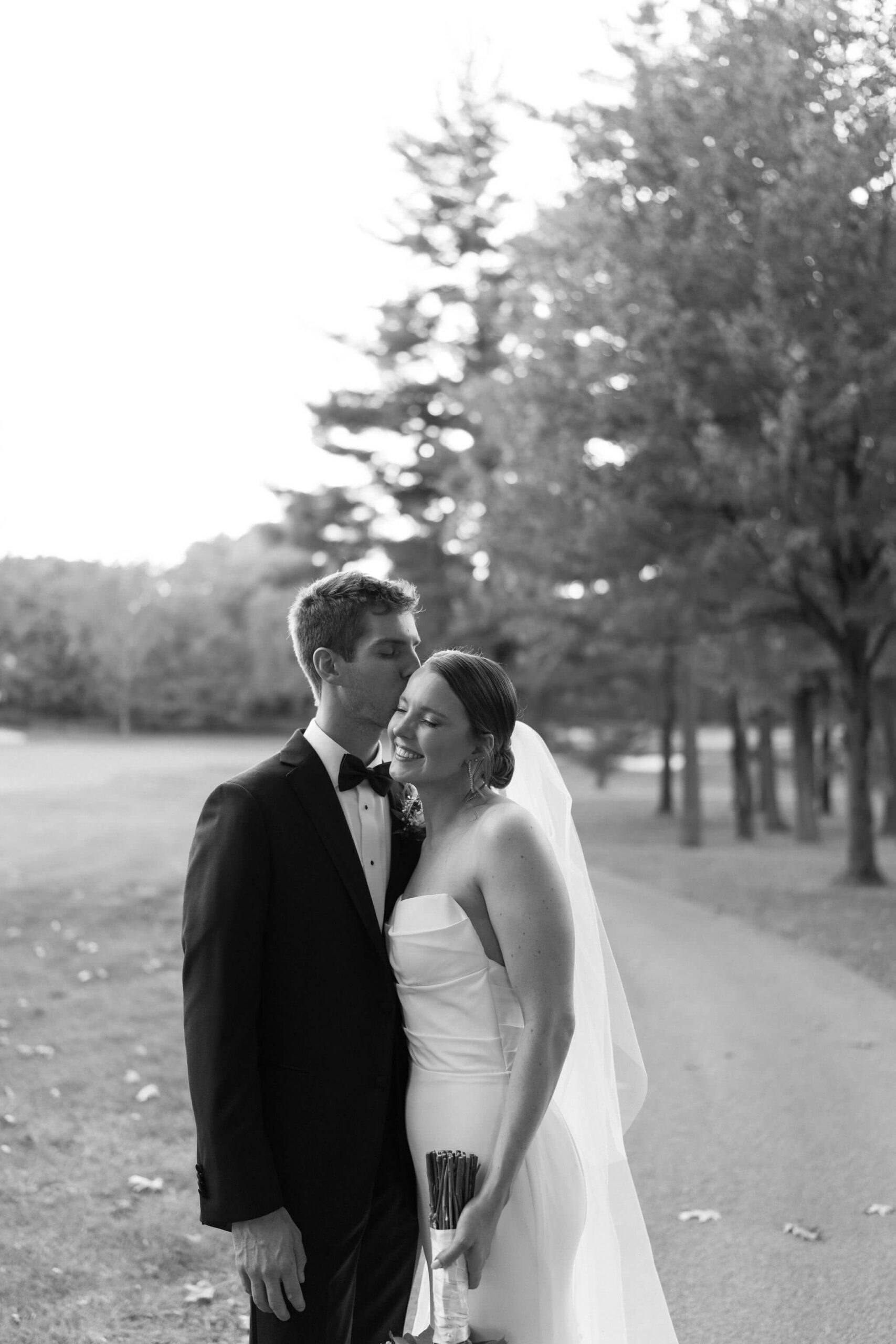black and white image of groom kissing bride on the cheek on a path through tall trees