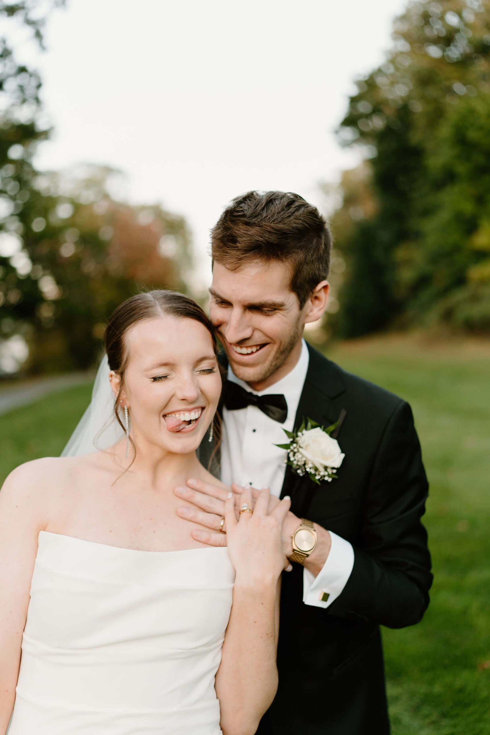 bride sticking her tongue out, groom laughing toward her behind her, both of their left hands on her left shoulder to show off their new wedding rings