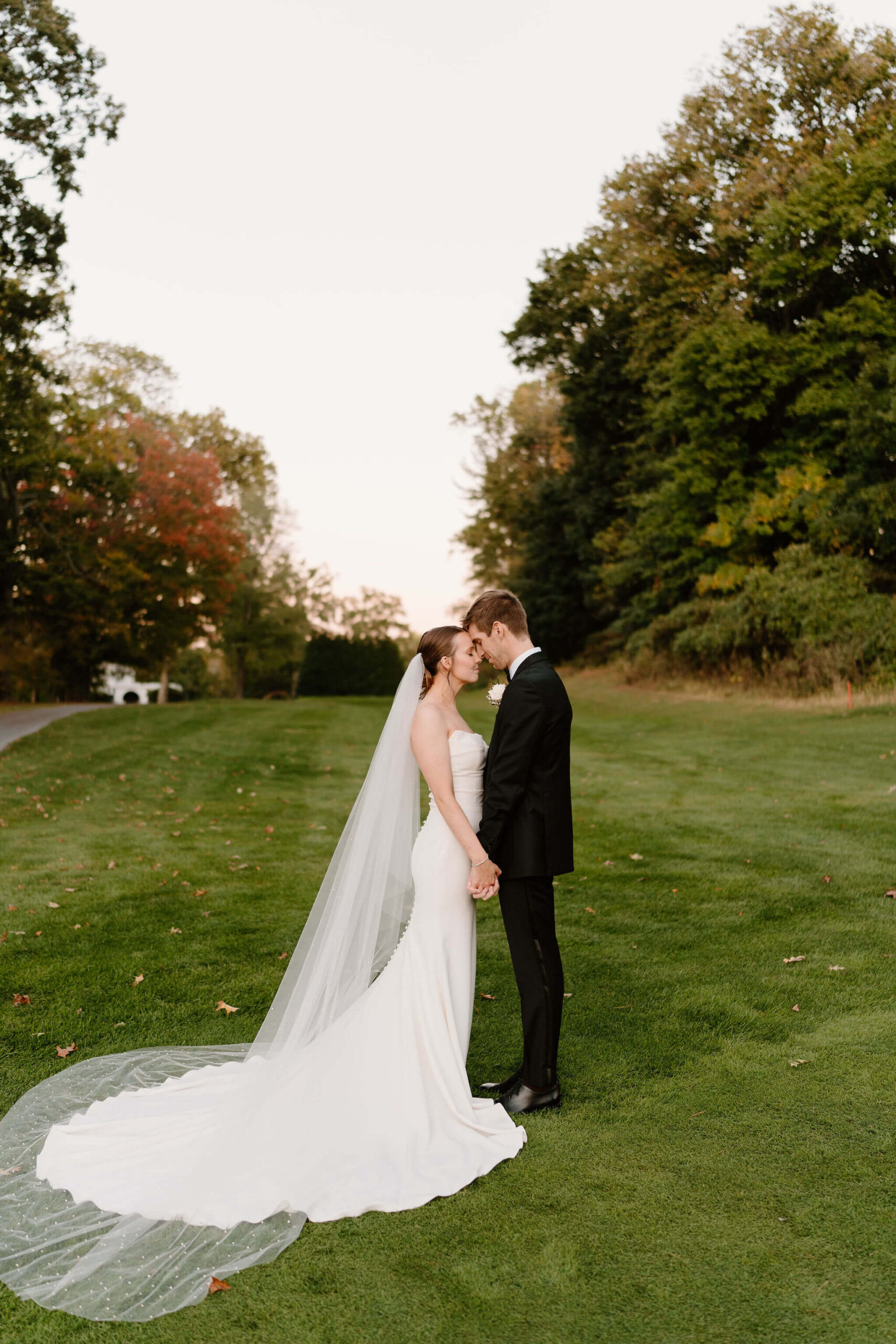 bride and groom holding hands and pressing their foreheads together, eyes closed, while the bride's train and veil lay out behind her. The sunset makes the leaves and grass around them glow with golden hour light.