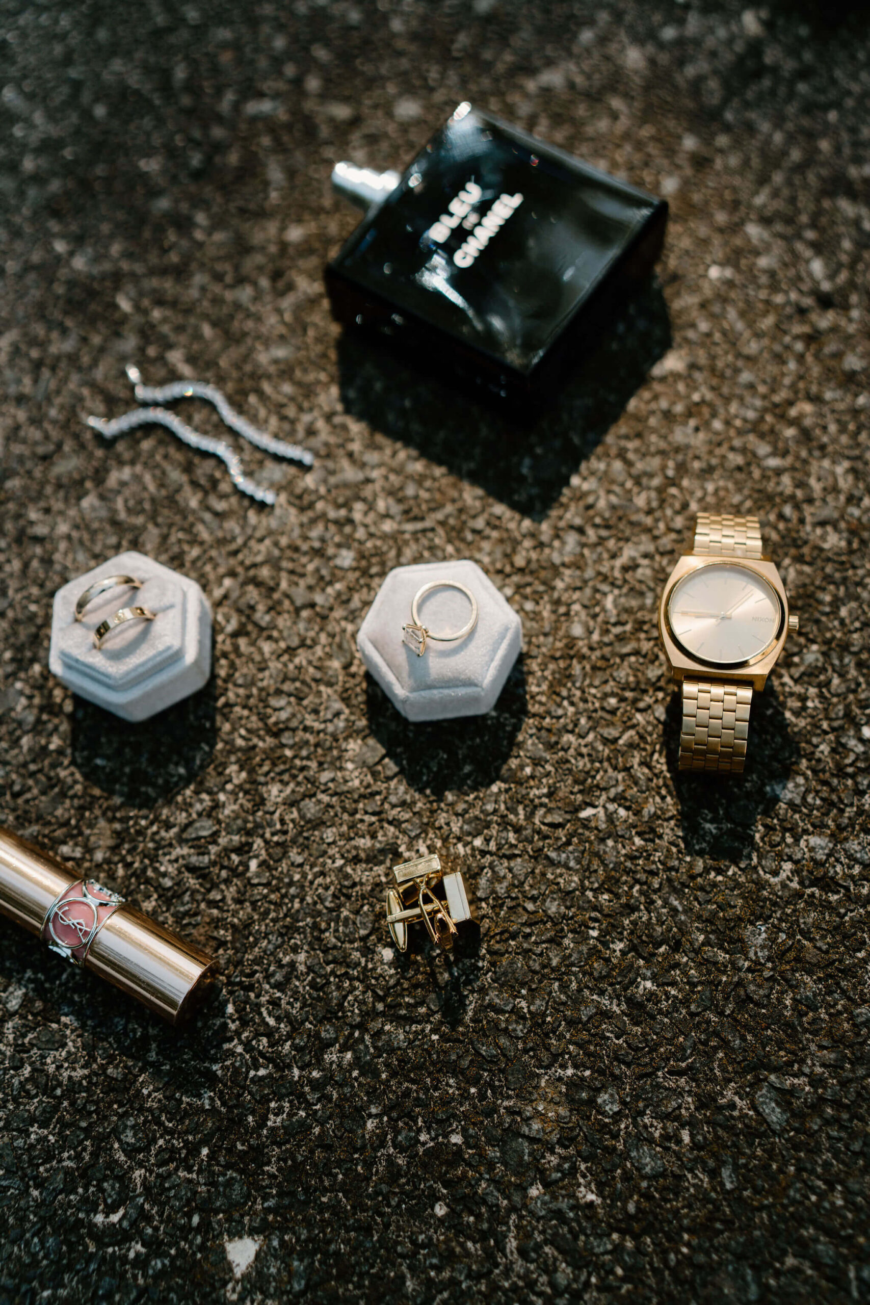 flat lay of an engagement ring, wedding bands, lipstick, cuff links, watch, and cologne