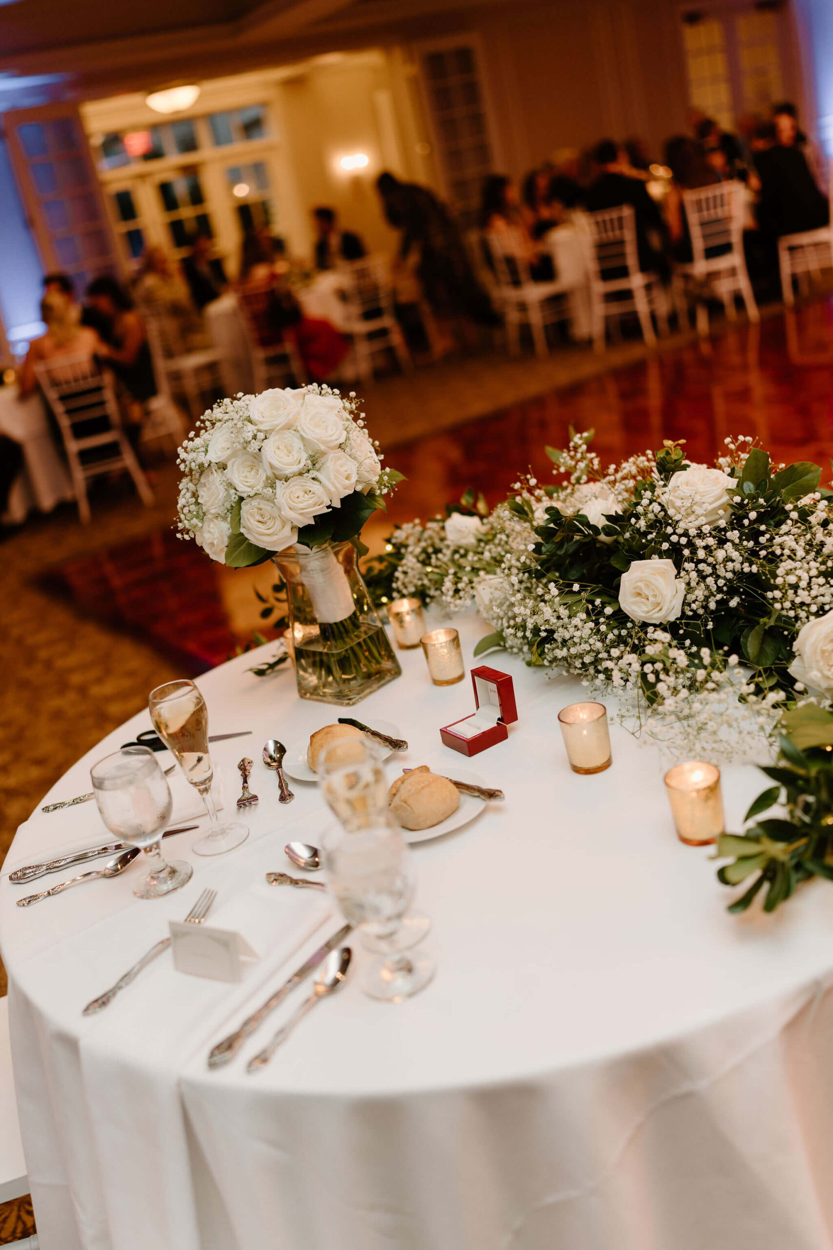 sweetheart table in a wedding reception, with a ring box open on the white linen tablecloth and a bridal bouquet of white roses in a vase