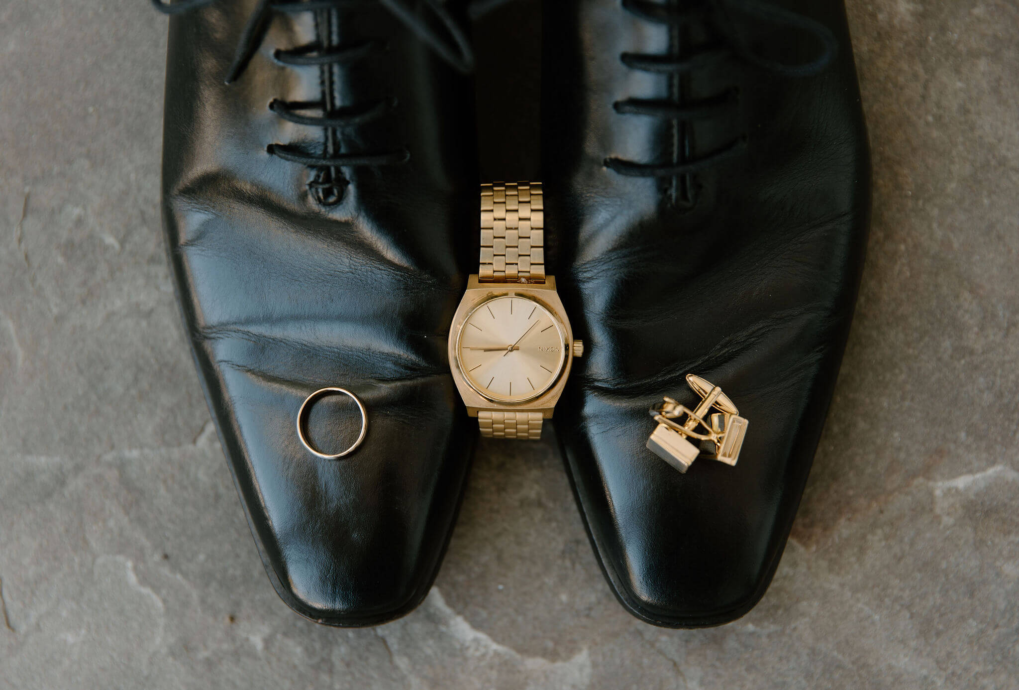 close up of a pair of black men's dress shoes with a gold watch propped in between them, a gold wedding band on the left shoe and two gold cuff links on the right shoe