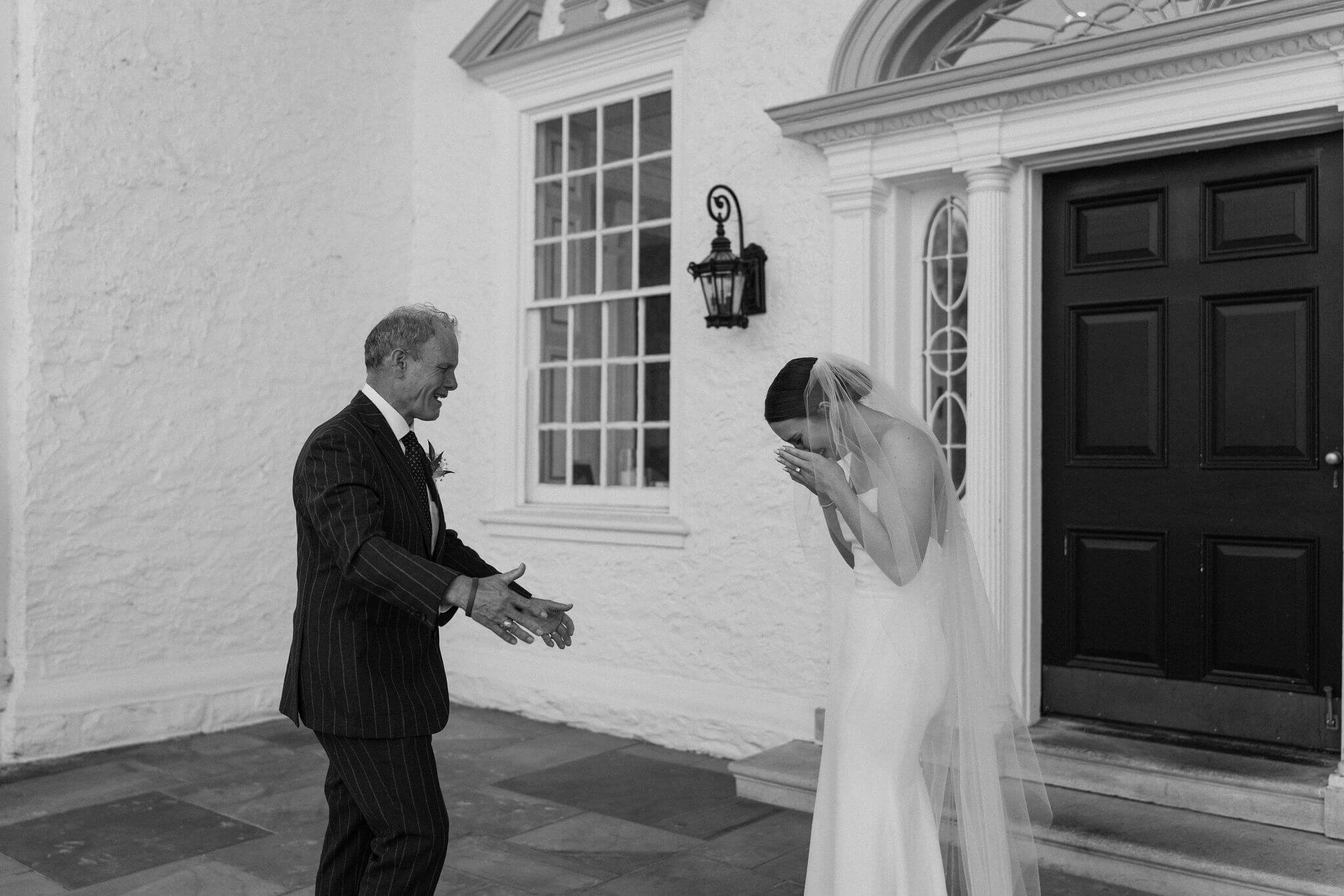 black and white image of a bride crying into her hands while her father walks toward her with his arms open after their first look
