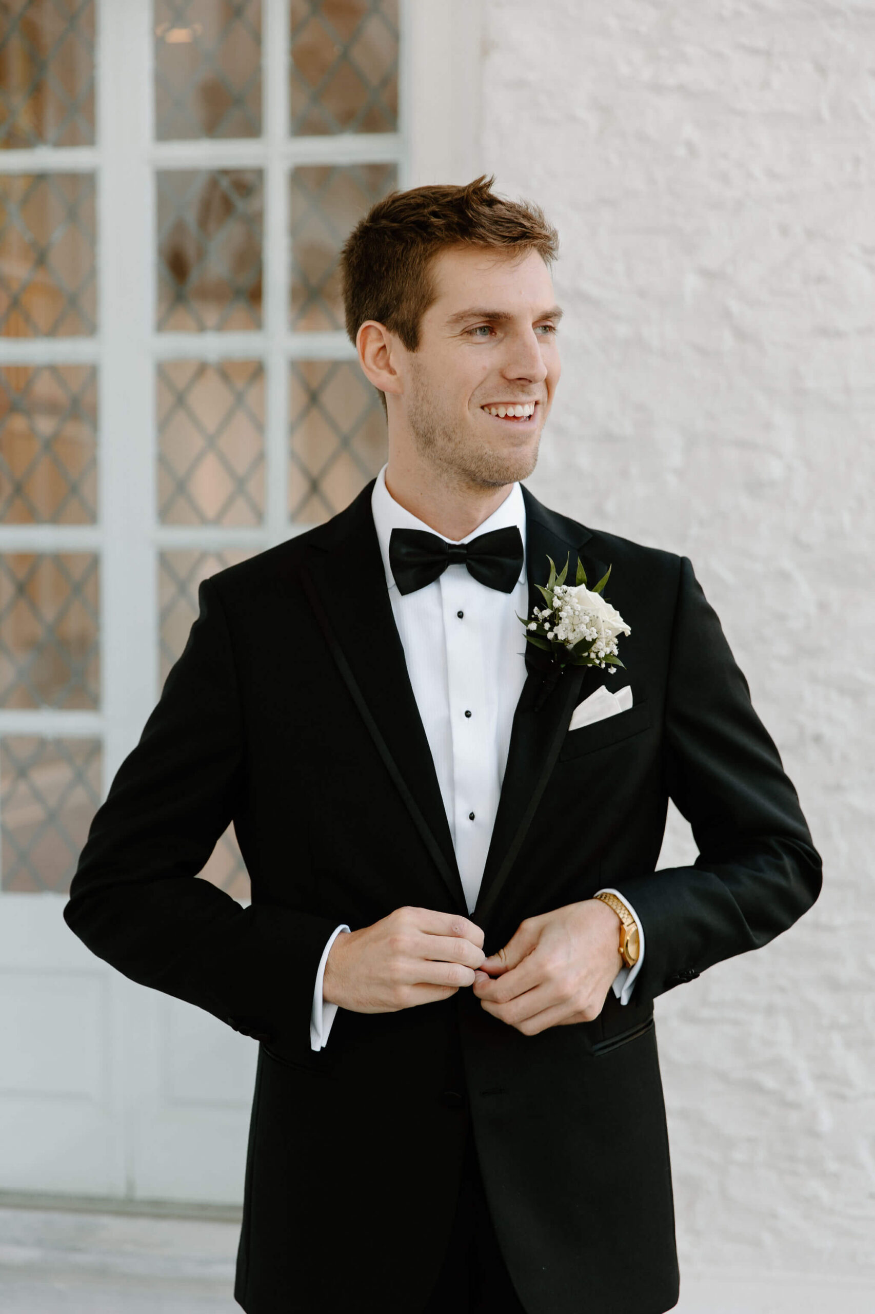groom in a black suit with a boutonniere buttoning his jacket while smiling off camera