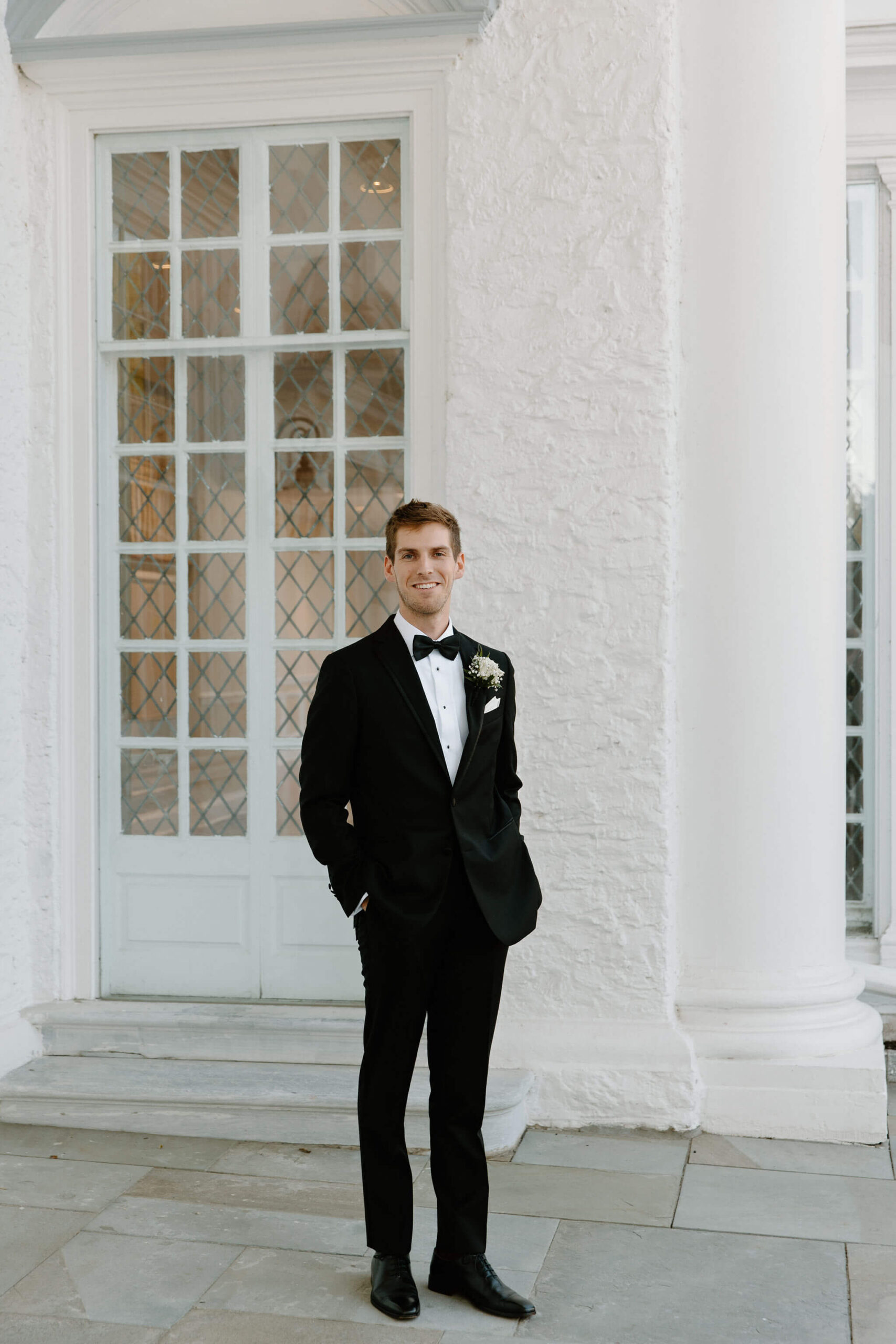 Groom in a black suit smiling at the camera with his hands in his pockets in front of the clubhouse at Radnor Valley Country Club