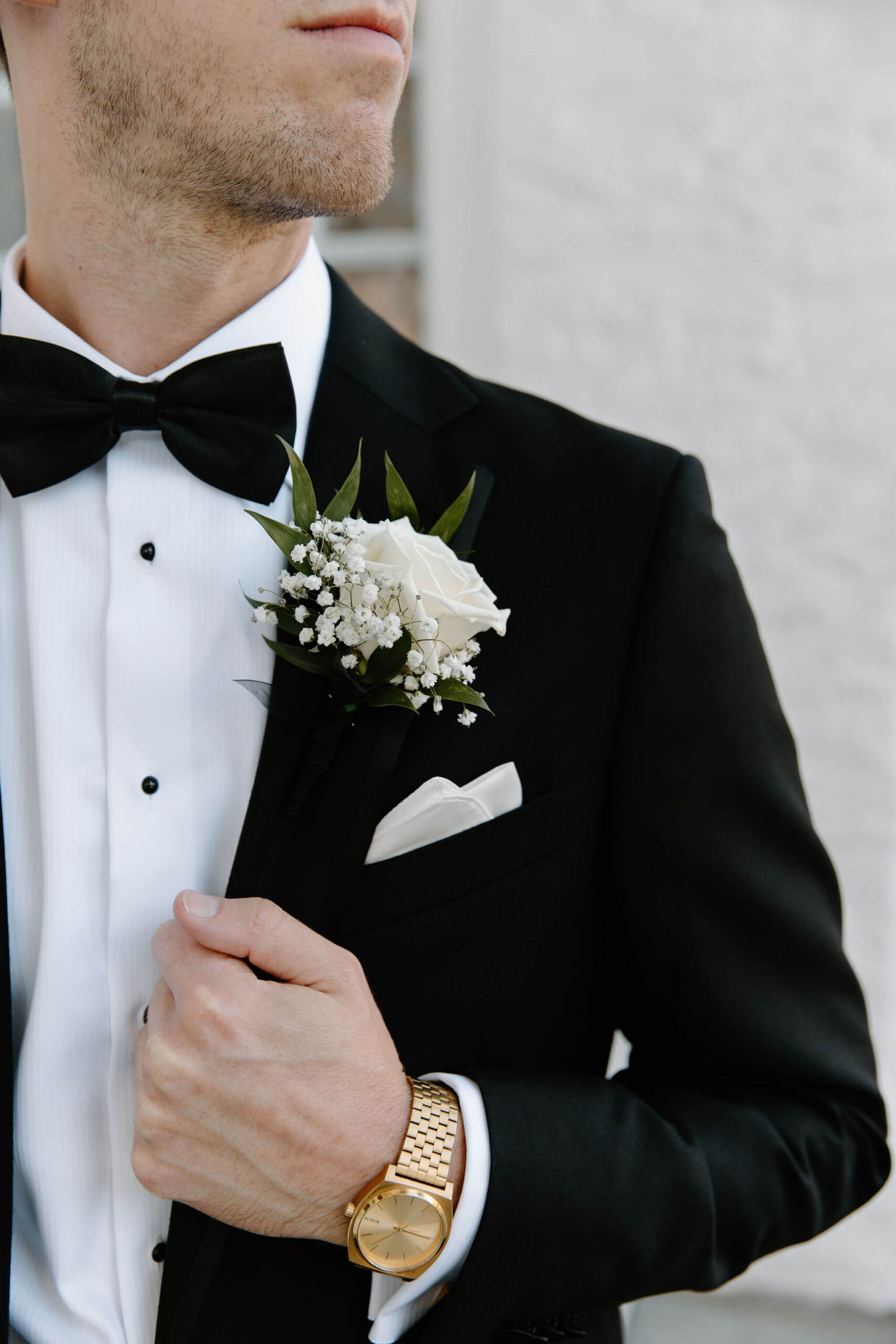close up of groom looking off camera, holding his jacket lapel with his left hand, sporting a gold watch and white rose boutonniere