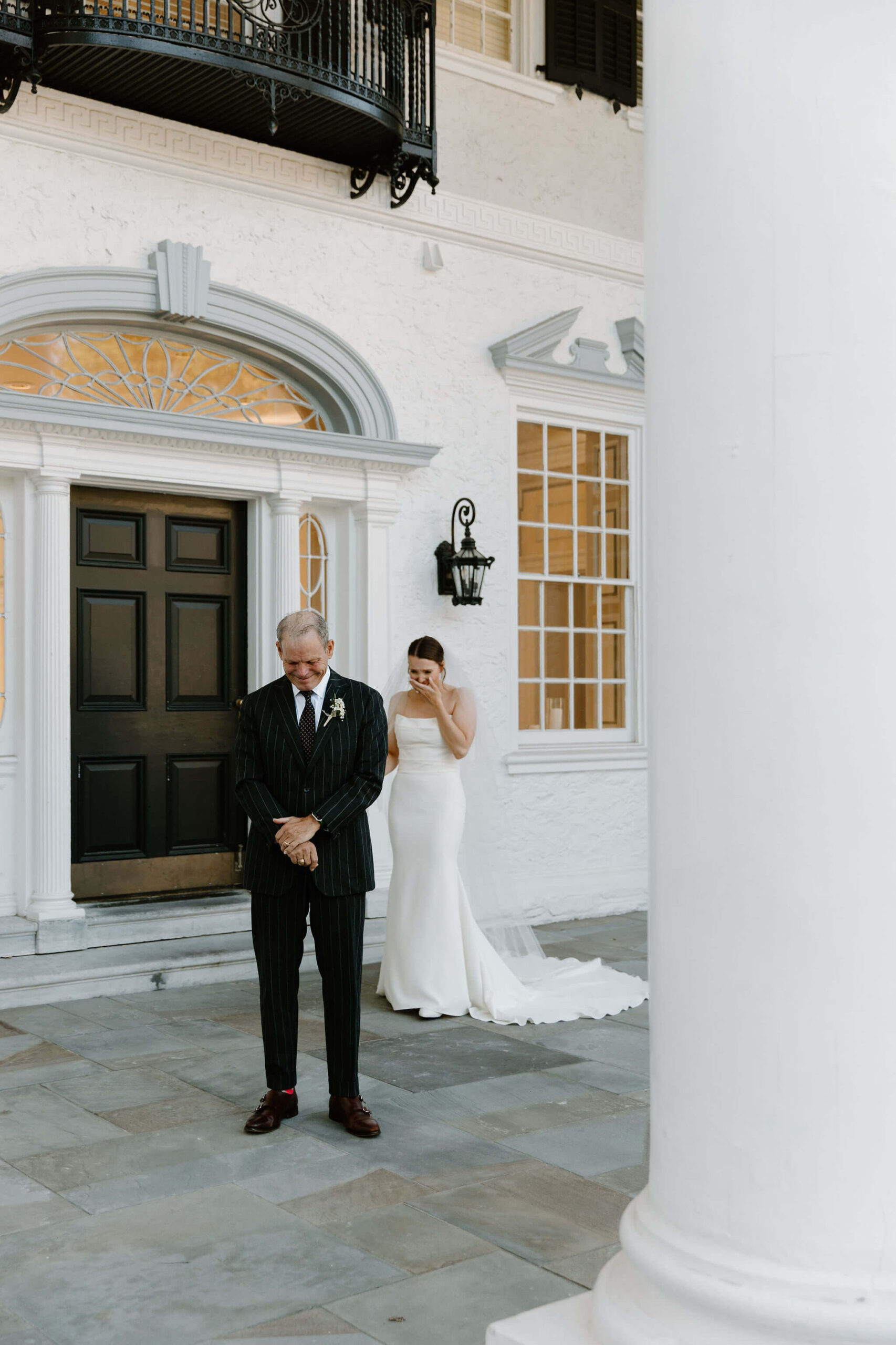 bride in a white wedding dress with a train and a floor length veil hunched over and crying before her first look with her father, standing with his back to her in a suit.