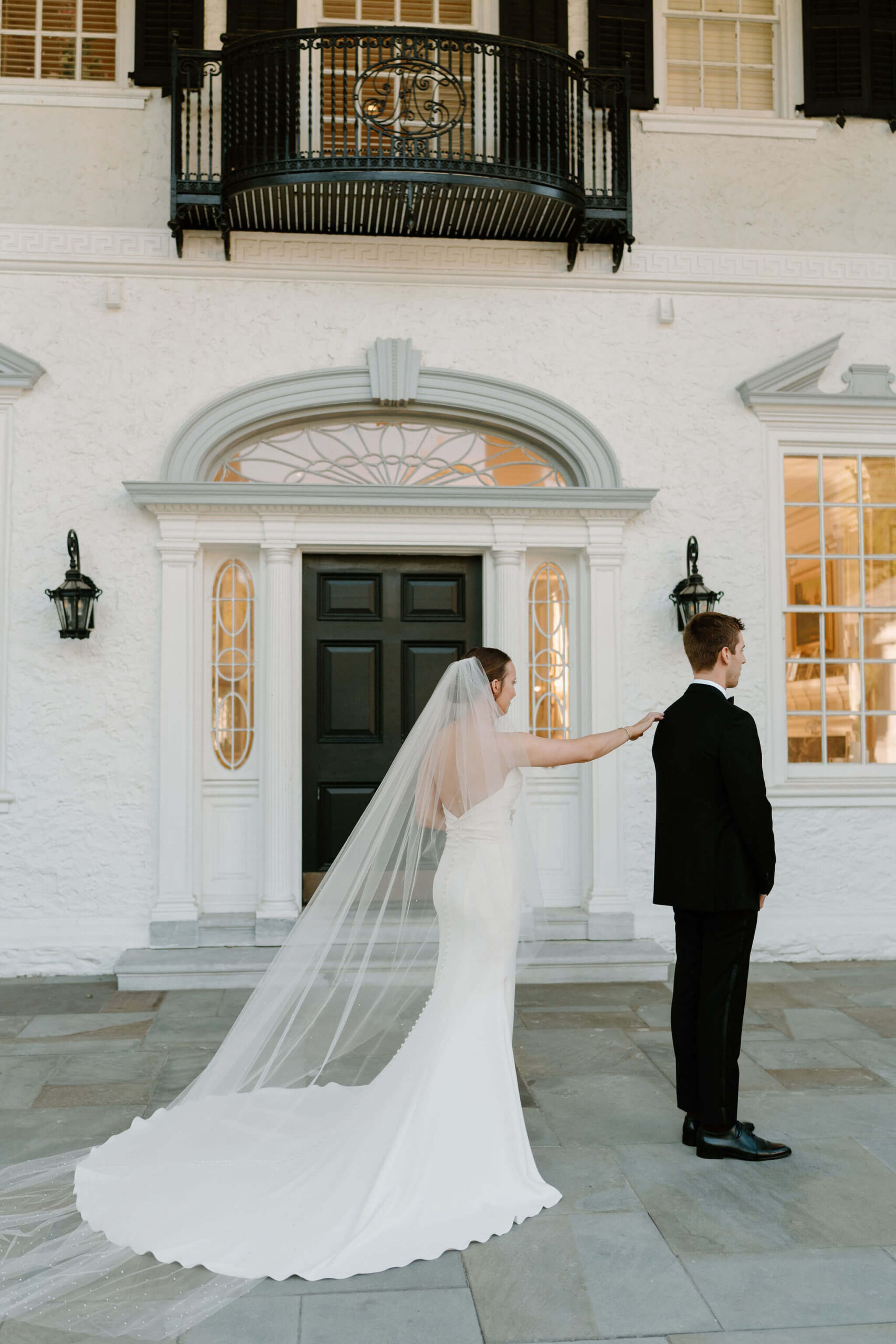 bride (long white open-back wedding dress and cathedral length veil) tapping her groom (black suit) on the shoulder for their first look