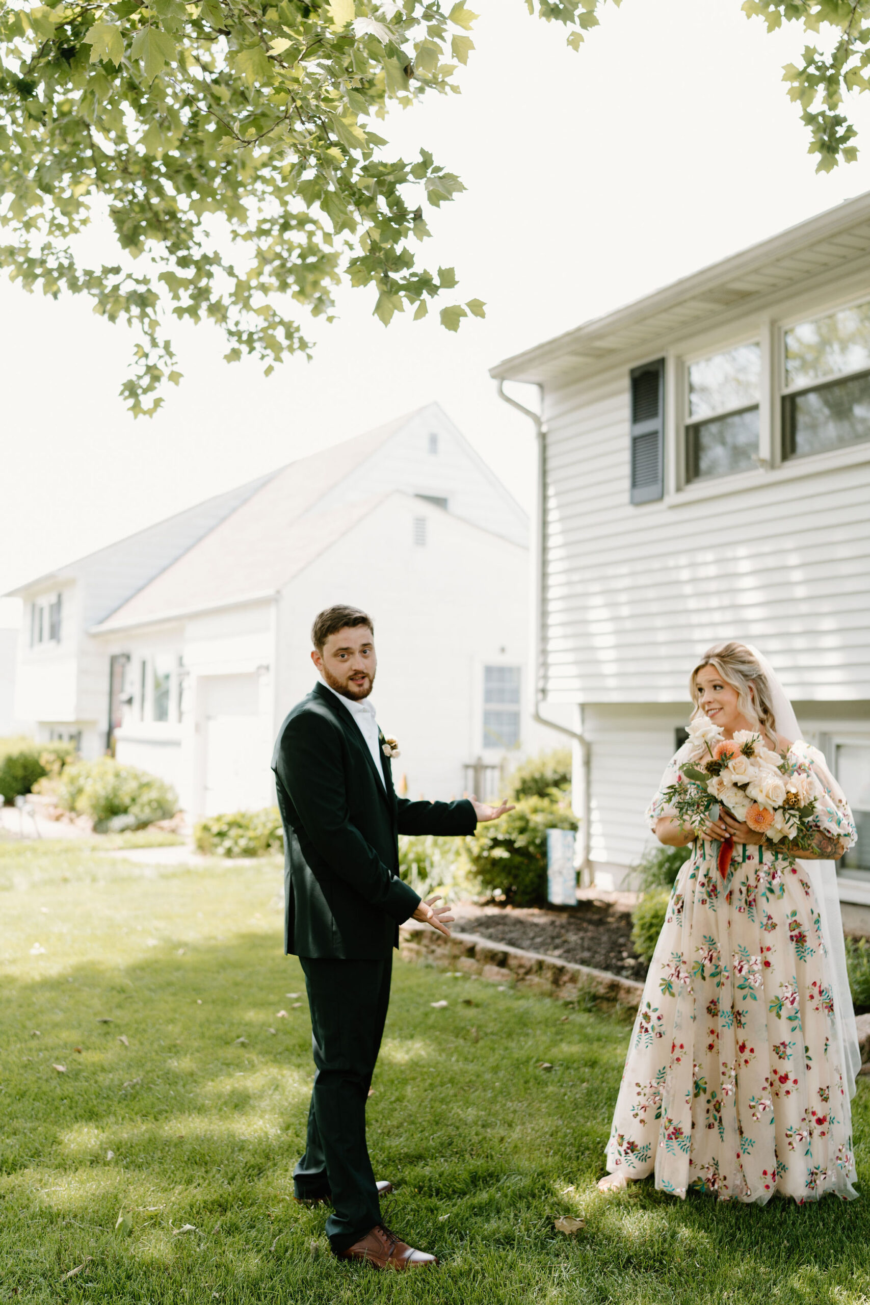 groom in a dark green suit looking at the camera and gesturing toward his bride in appreciation while she smiles at him, holding her bouquet and wearing a colorful alternative wedding dress