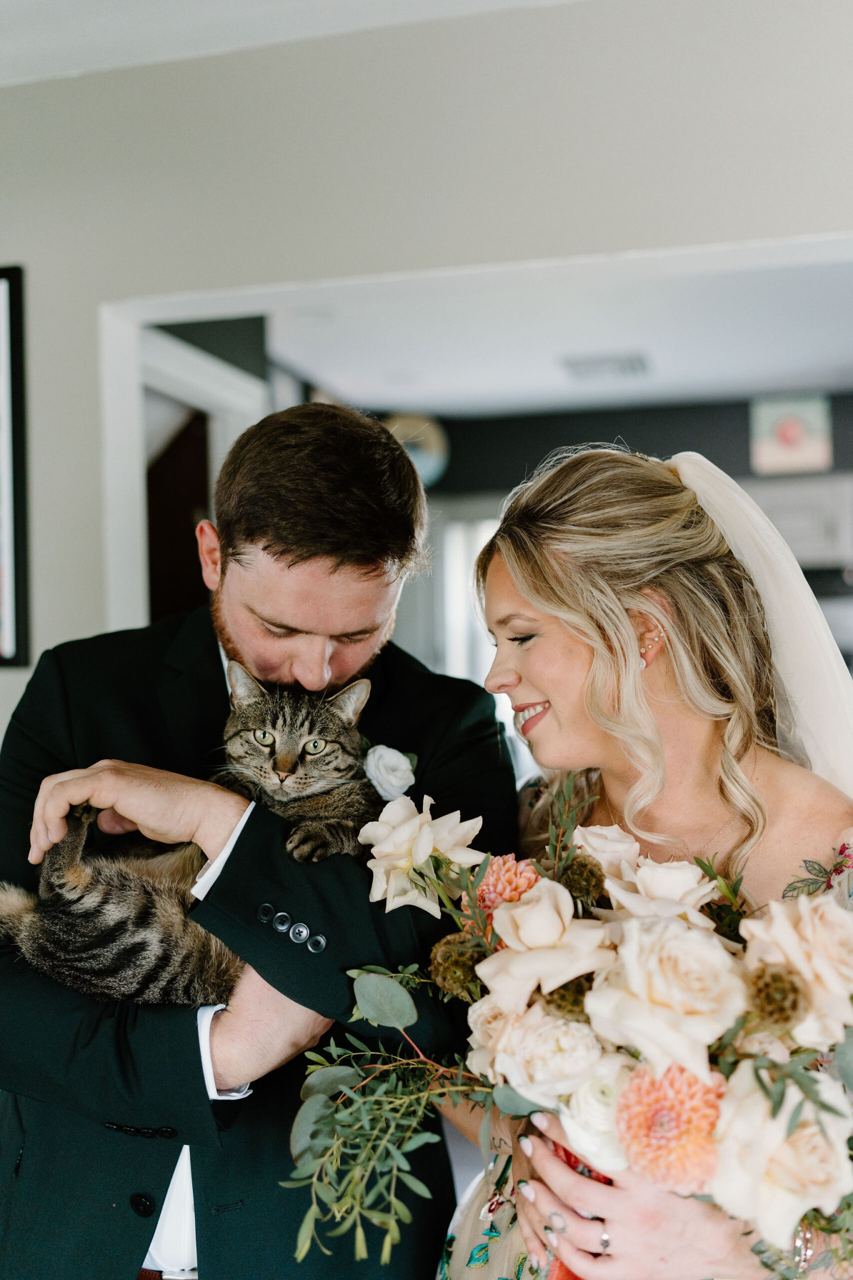 bride and groom holding their gray tabby cat, kissing him on the head and smiling at him.