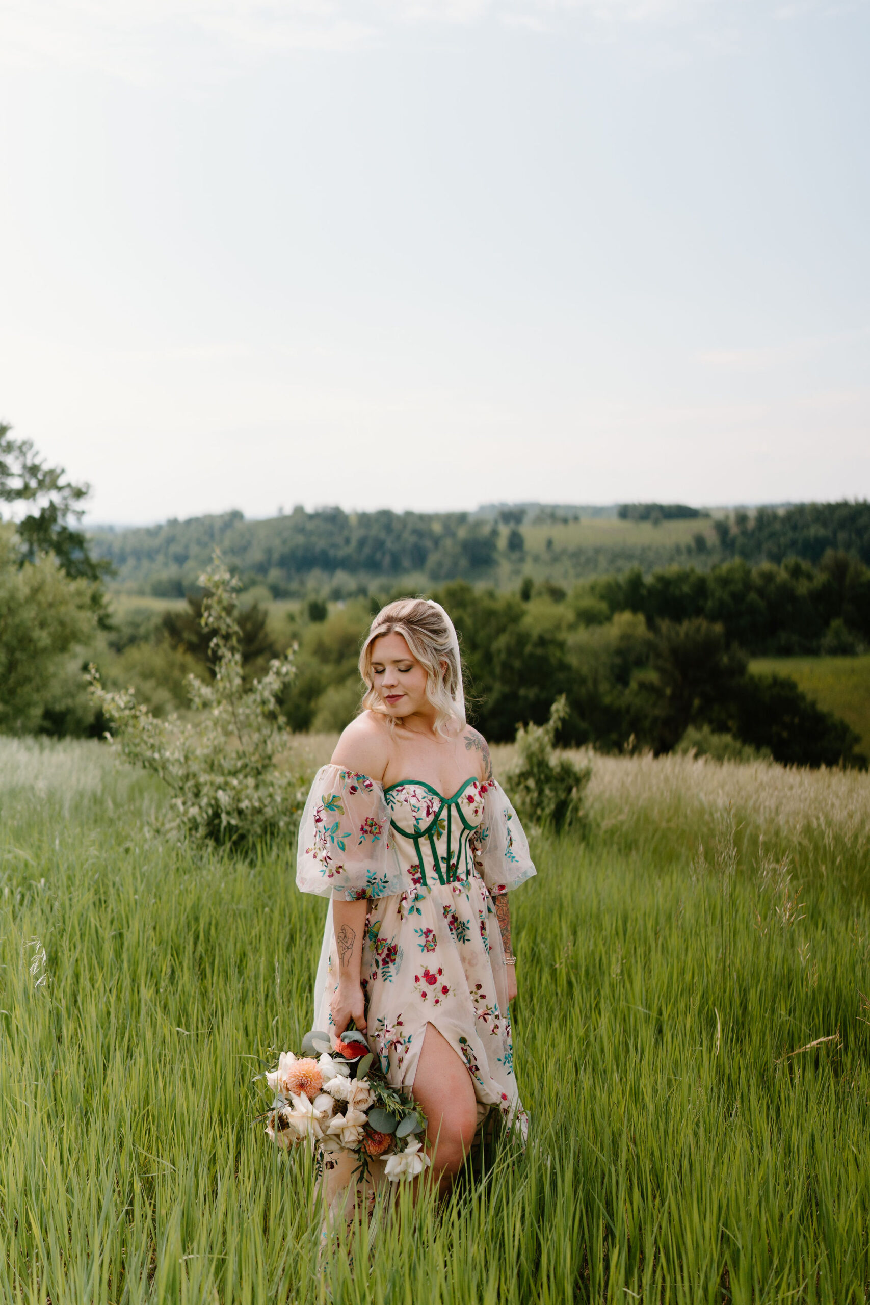 bride in a colorfully embroidered wedding gown with green boning smiling softly over her shoulder and holding a bouquet down at her side, standing in a field of tall grass