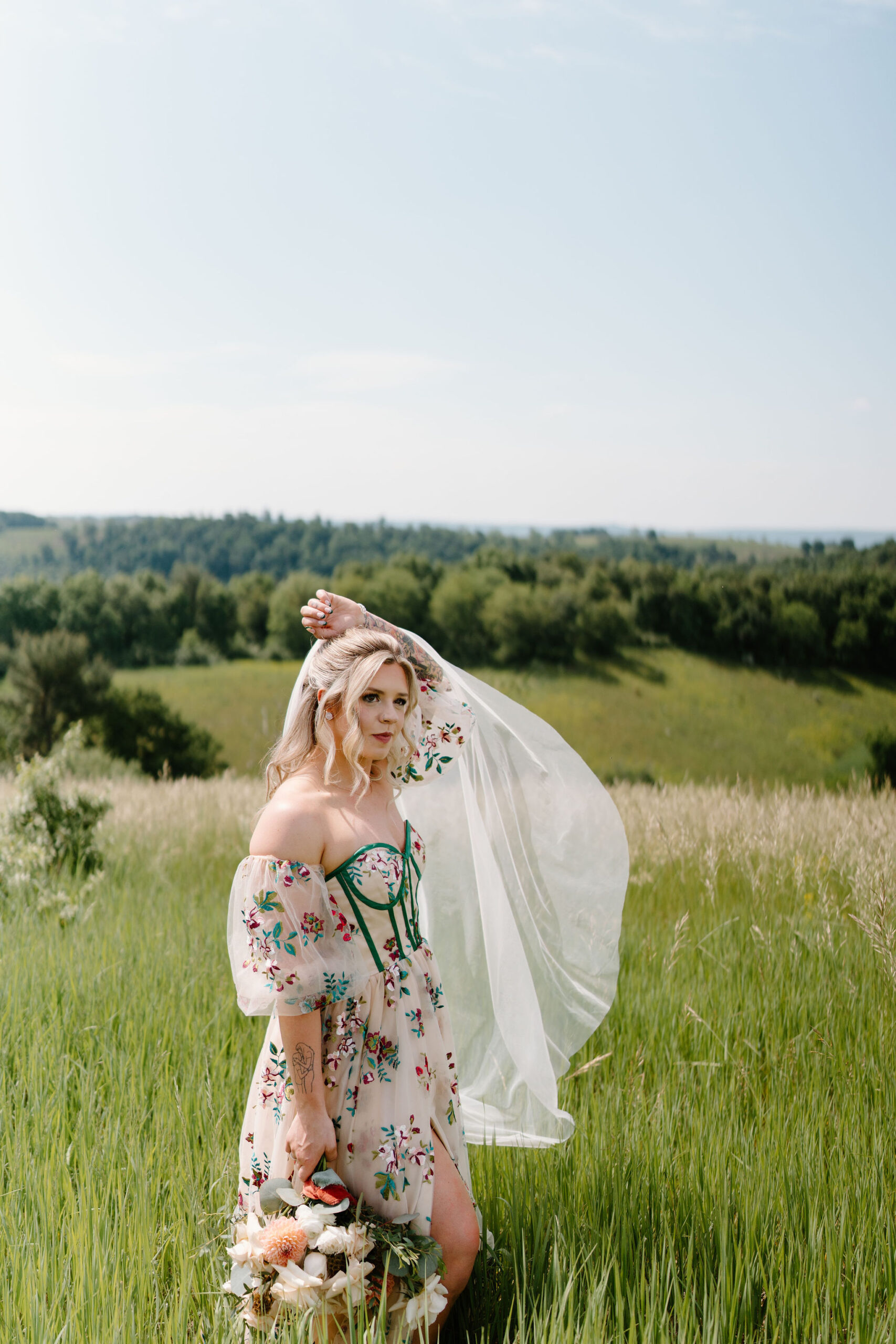 alternative bridal look with tattoos and a colorfully embroidered wedding dress, tossing her veil over her head with one hand and holding her bouquet of flowers down at her side