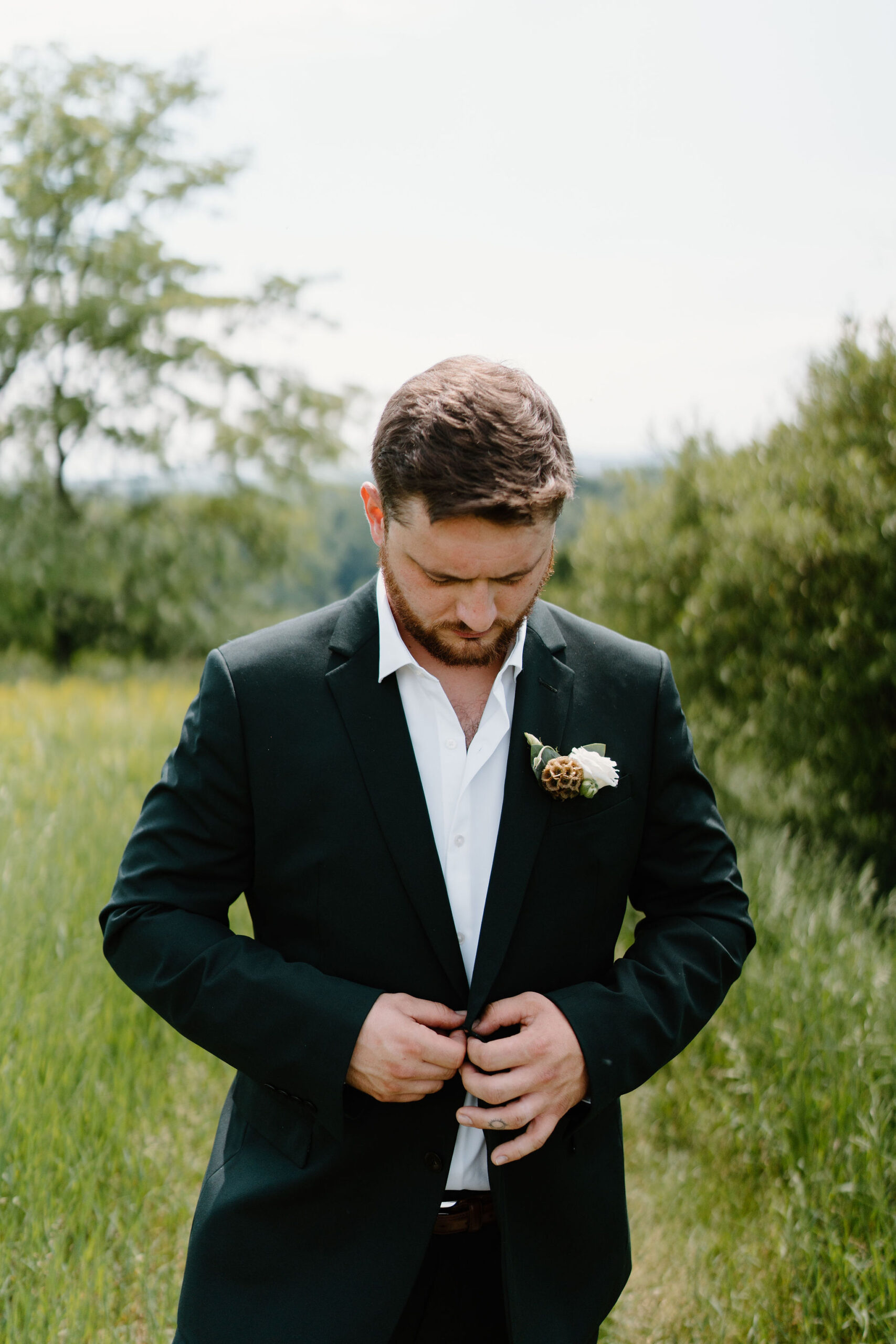 groom (green suit, boutonniere) buttoning his suit jacket in a field