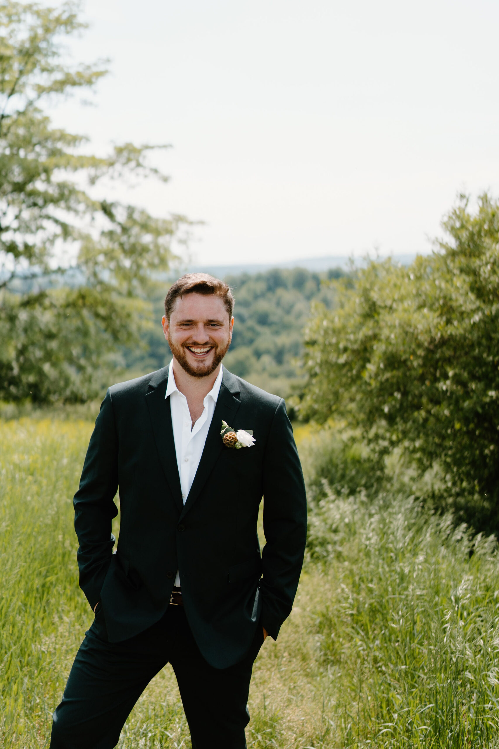 groom (dark green suit, boutonniere) smiling at the camera in a field with rolling hills behind him, with his hands in his pockets