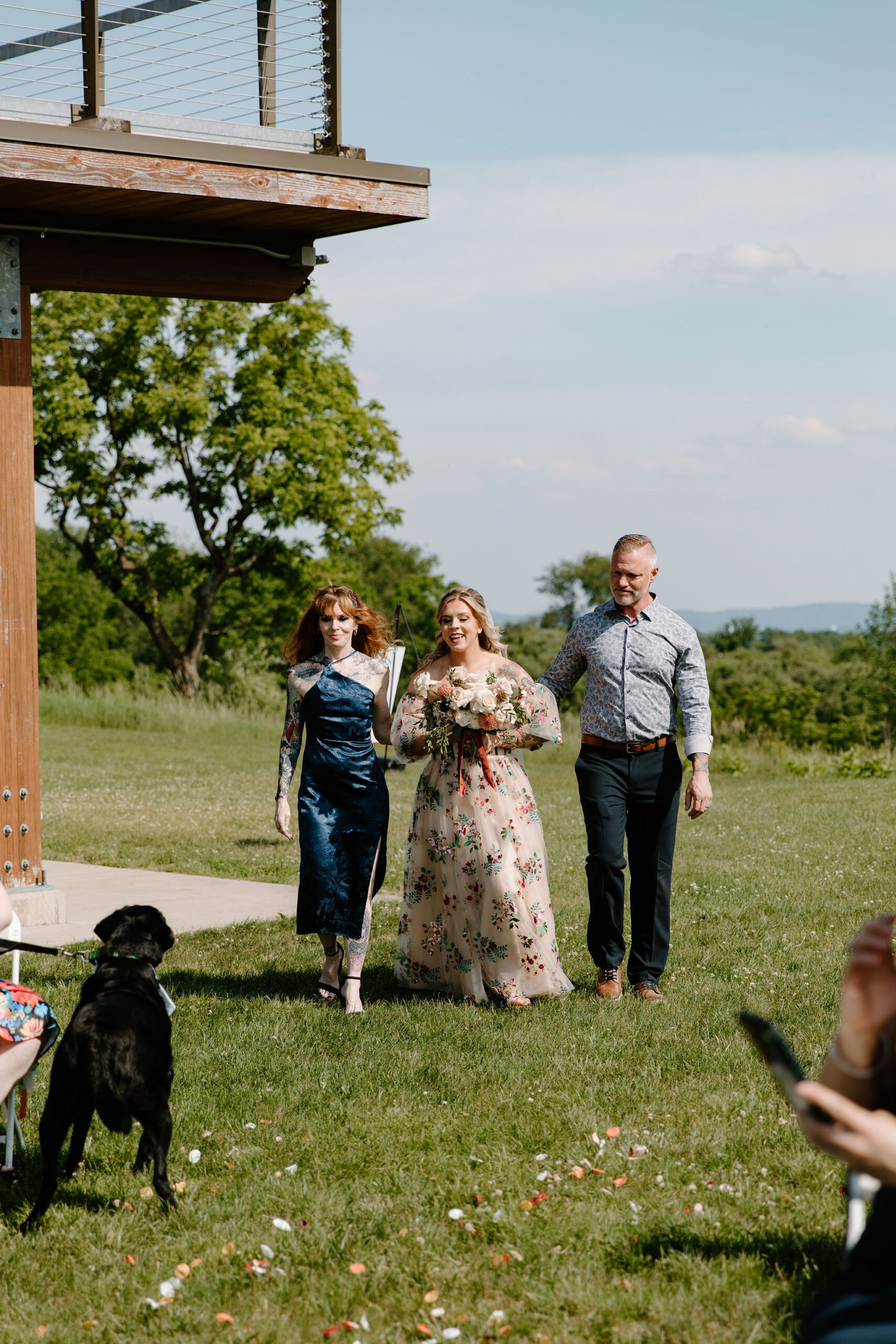 bride in an alternative, colorful wedding dress, being walked down the aisle by her parents while her dog tries to get to her