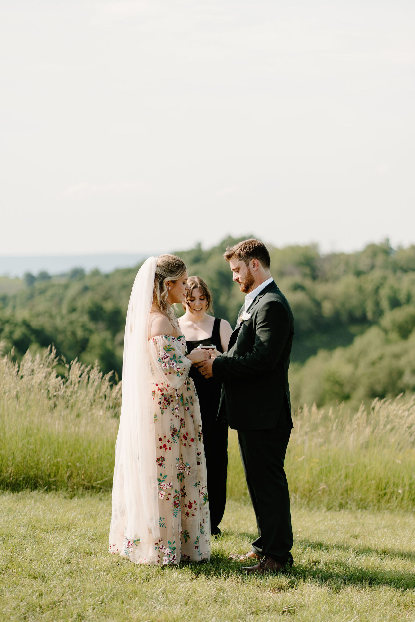 bride (brightly colored embroidery in her dress, and tattoos over her arms) holding groom's hands (dark green suit with boutonniere) during their wedding ceremony, their officiant standing behind them with rolling fields in the distance.