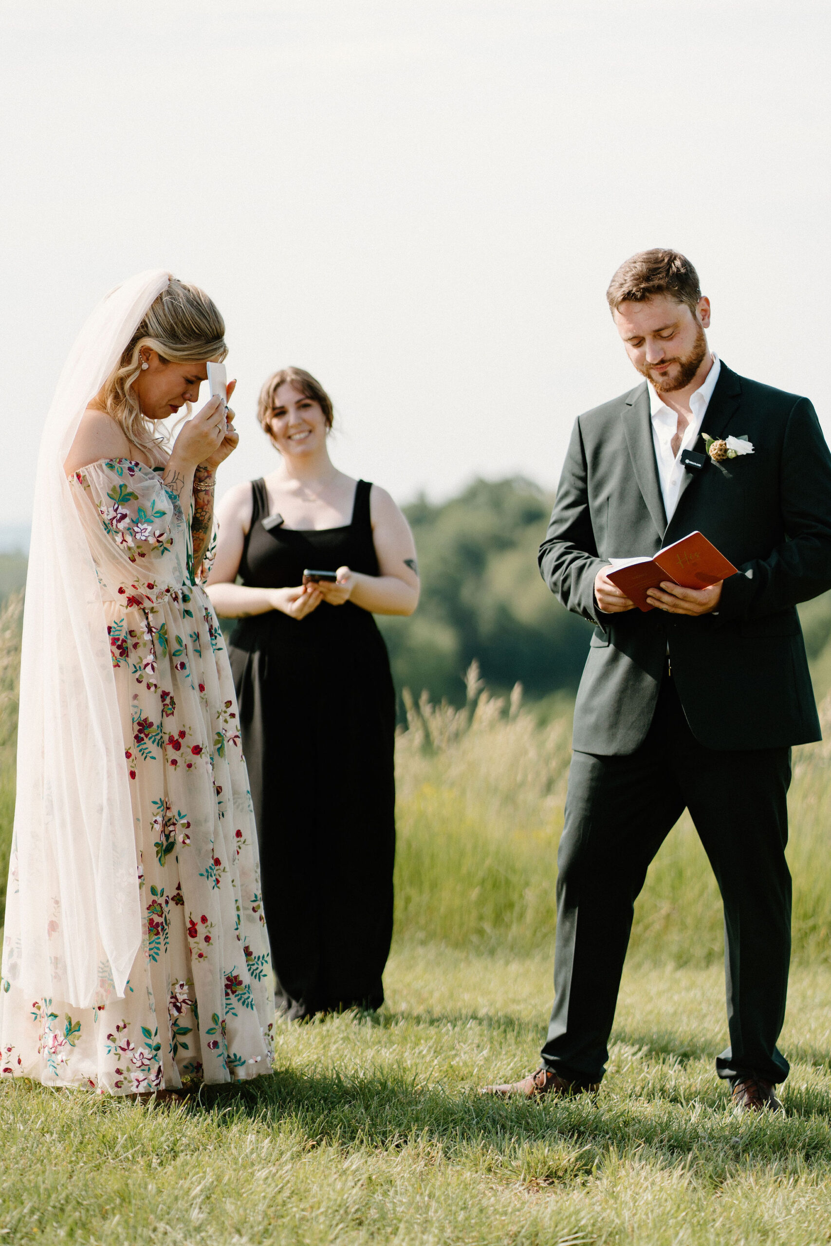 bride in a colorful wedding dress covering her face with her vow book and crying as her groom reads his vows to her, while their officiant smiles behind them