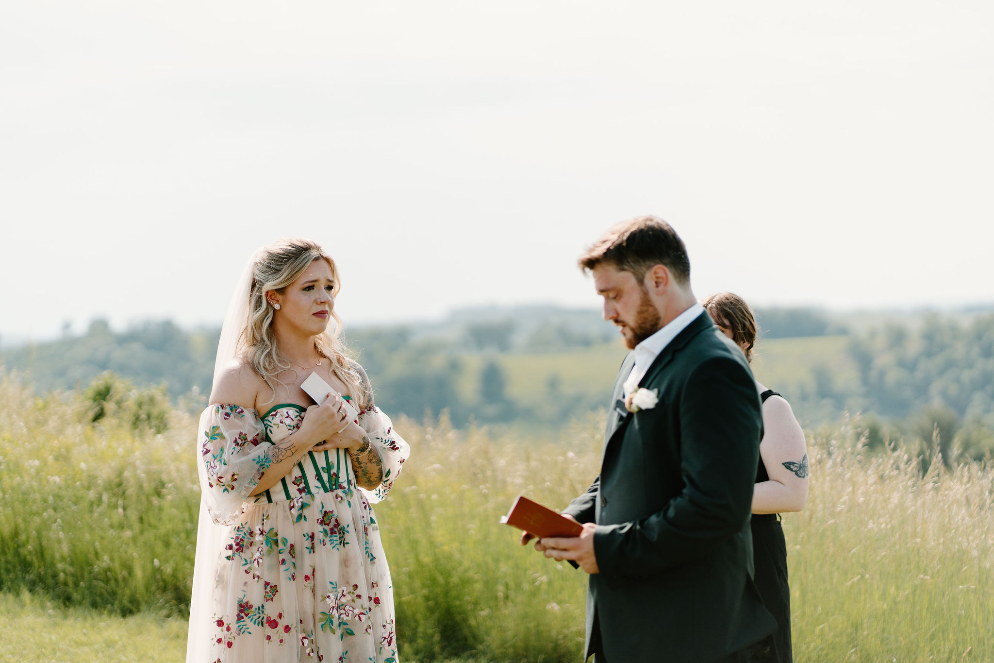 bride crying and holding her vow book to her chest, while her groom reads her his vows on a sunny day in a rolling green field