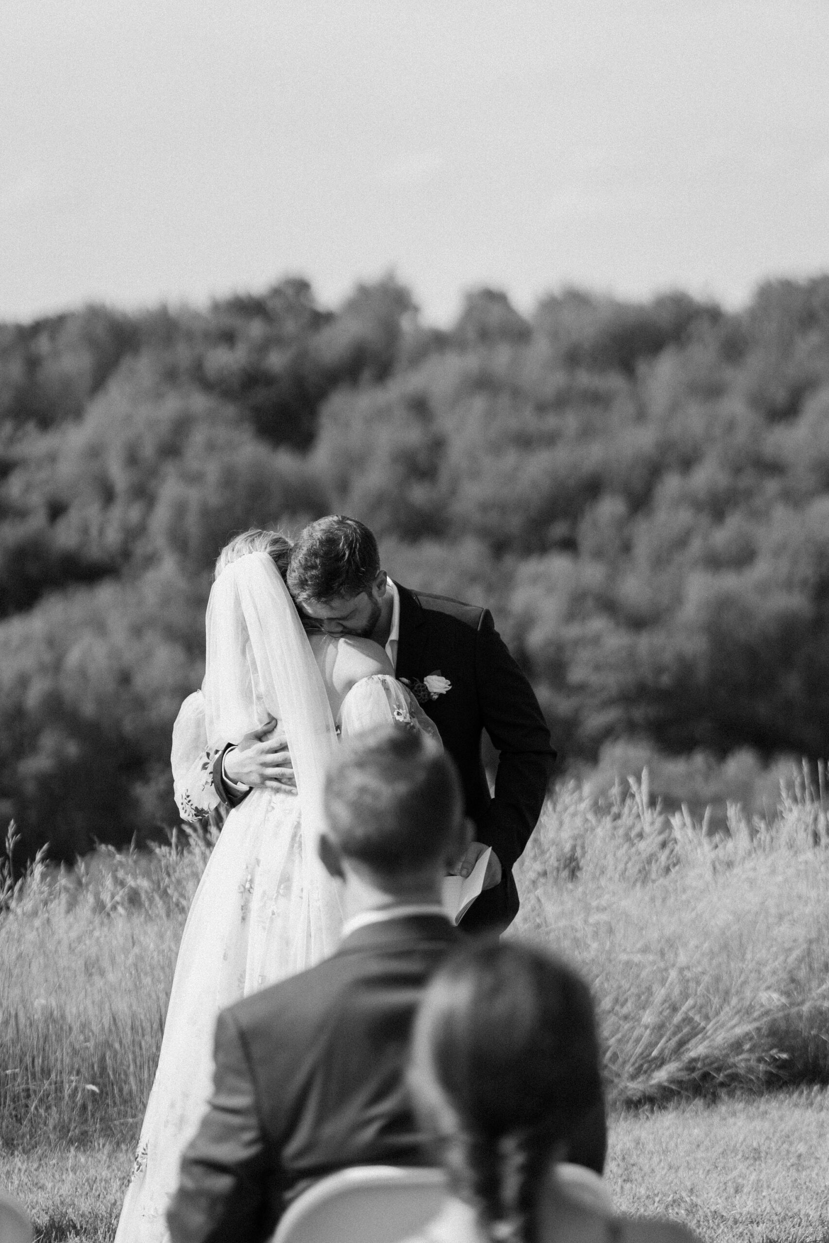 black and white image of groom holding his bride and kissing her shoulder during their wedding ceremony in between vow exchanges