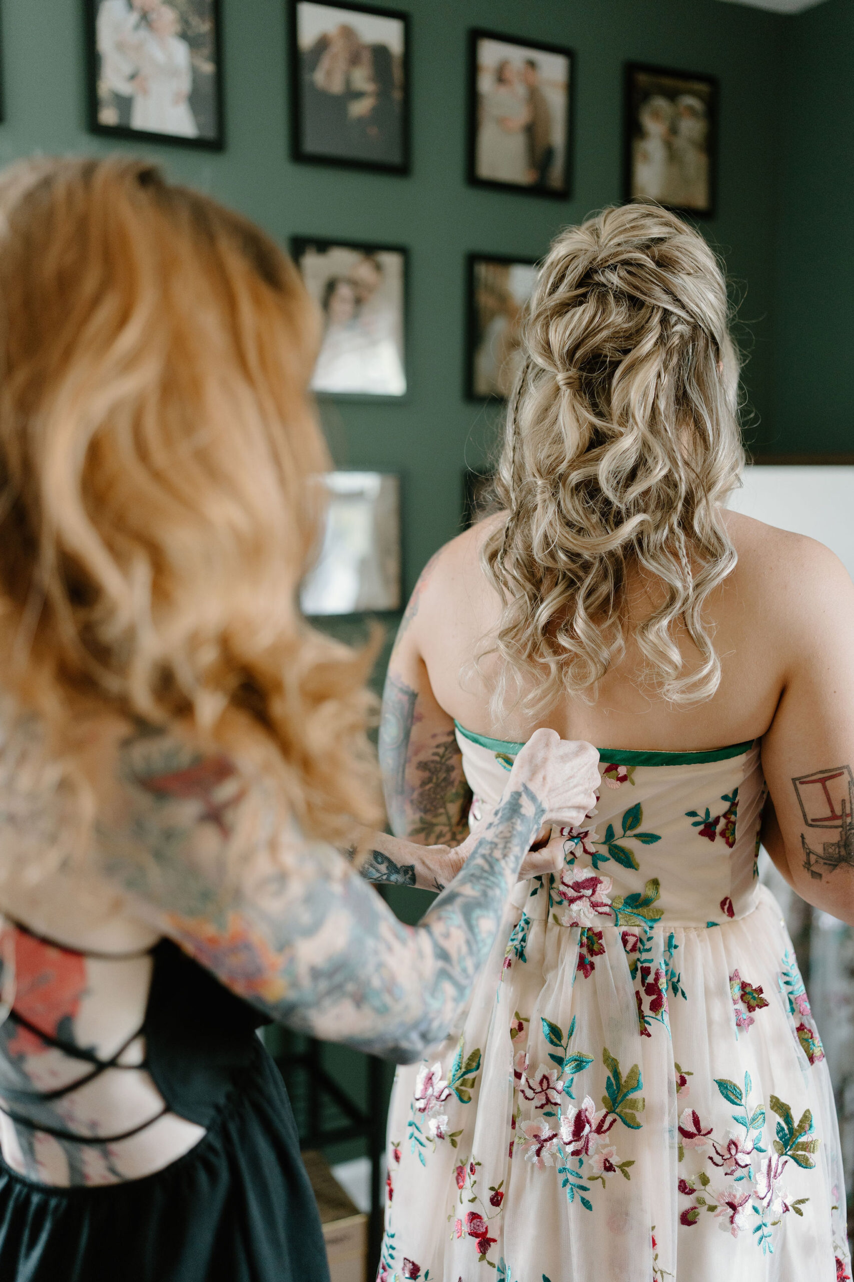 mother of the bride (red hair, heavily tattoed, black dress) helping the bride (blonde hair, tattoos) into her colorfully embroidered wedding dress