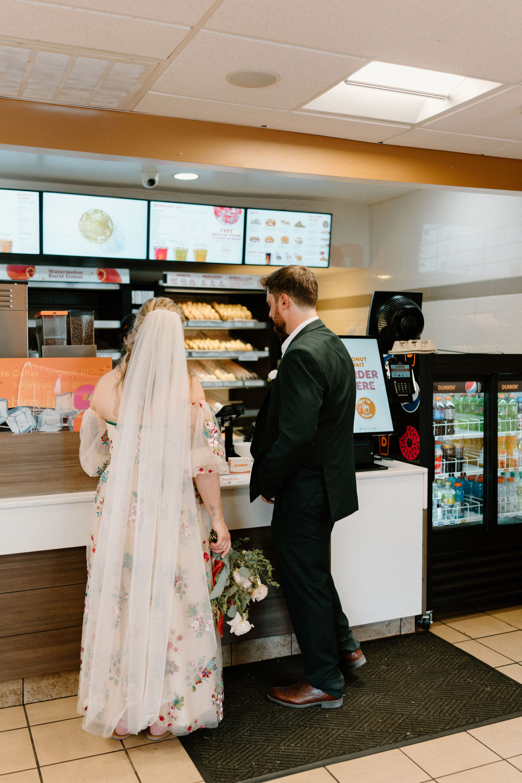 bride in a colorful wedding dress and groom in a dark green suit, placing an order at Dunkin Donuts at the counter