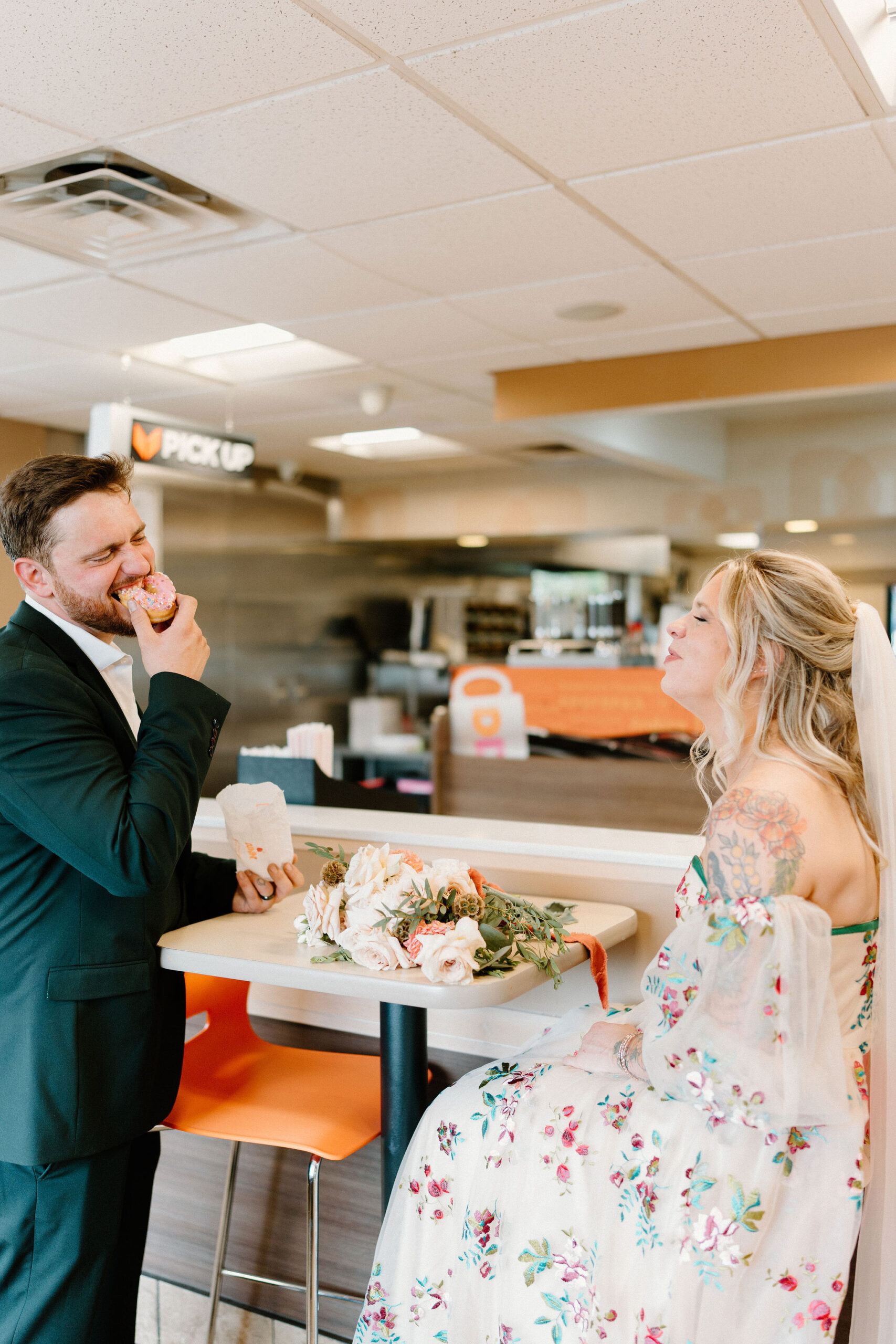groom taking a bite of a donut inside Dunkin Donuts while his bride laughs