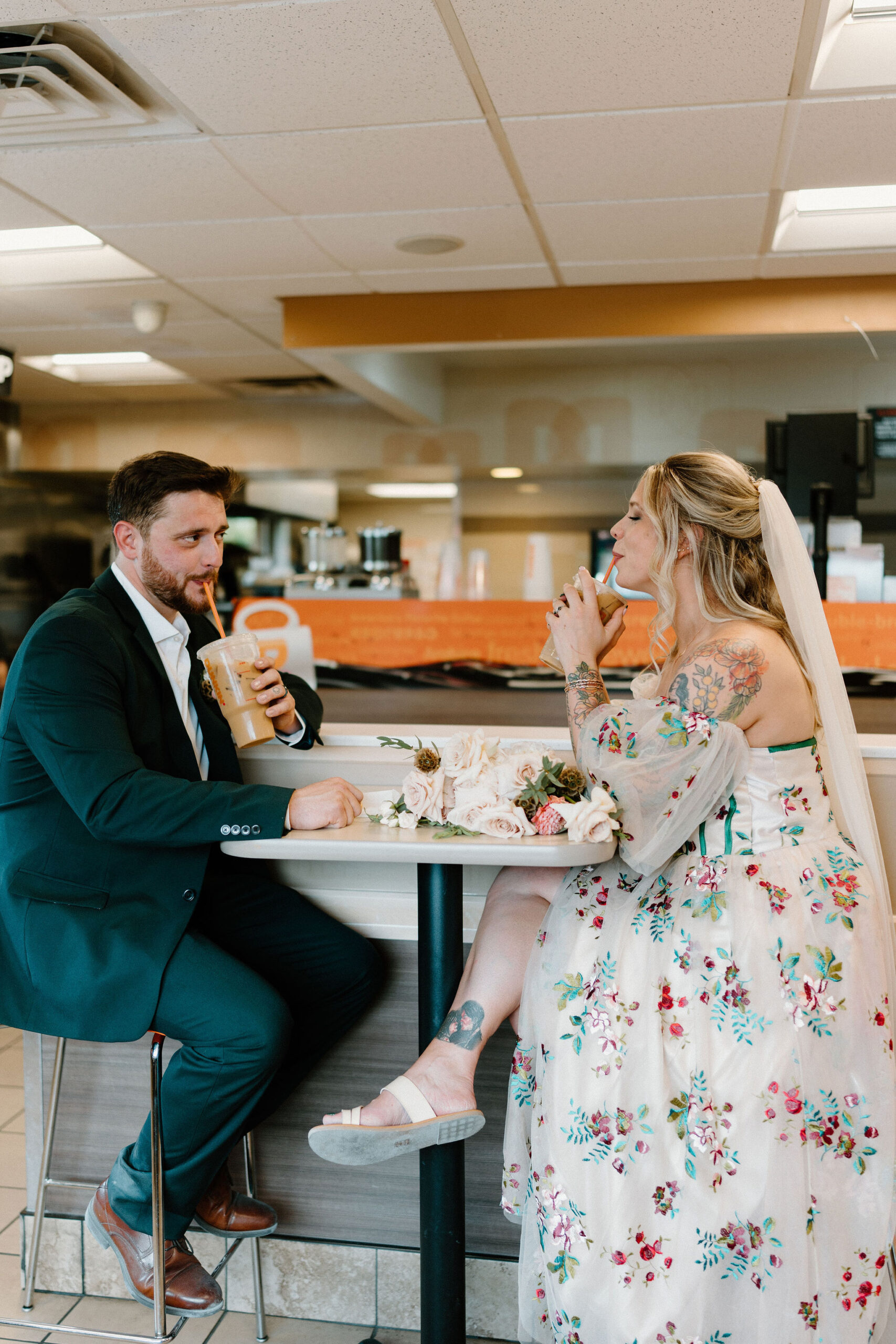 bride and groom sitting across from each other at a table in Dunkin Donuts, each taking a sip of their iced coffees