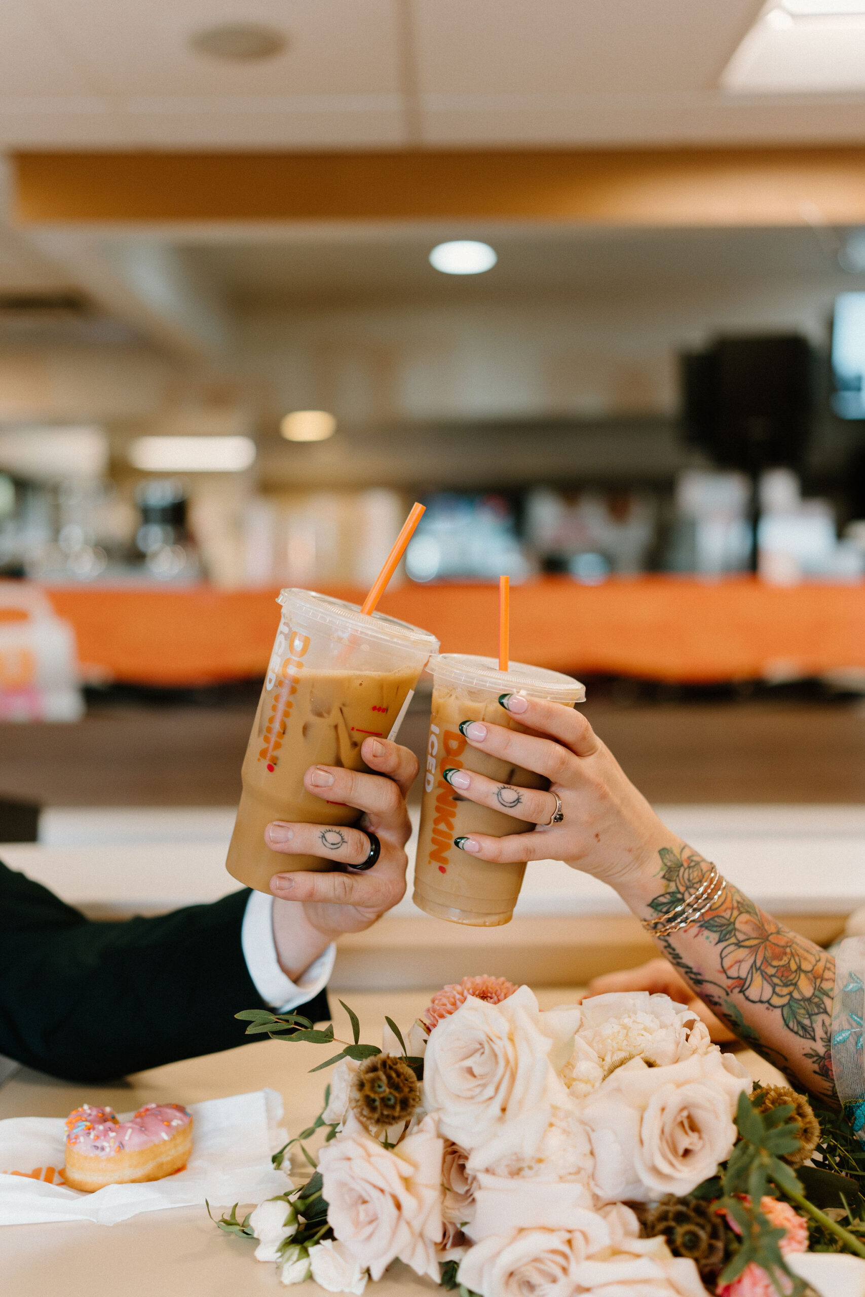 close up image of bride and groom's hands, both with wedding bands and a wedding tattoo on their ring fingers, toasting with their dunkin donuts iced coffees with a half eaten donut and her wedding bouquet between them