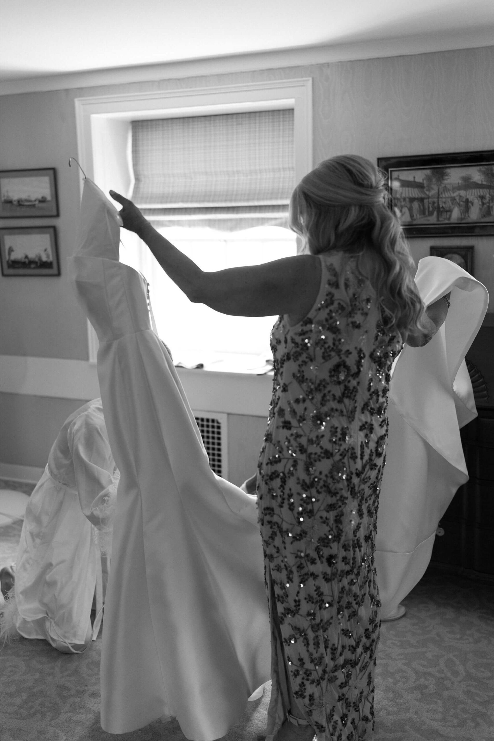 black and white image of the mother of the bride, holding the bride's wedding dress while the bride kneels to steam it