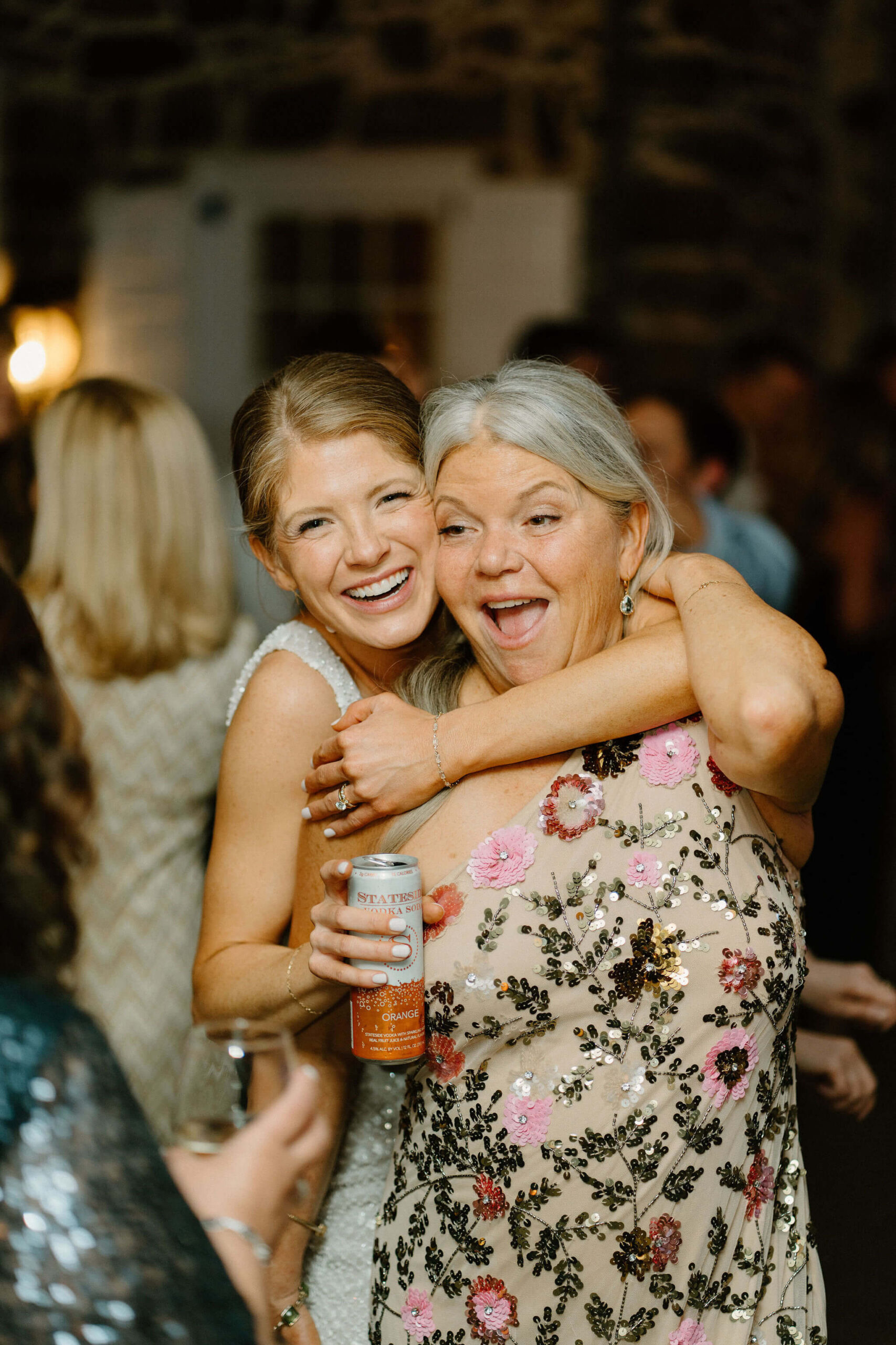 bride smiling with her arm thrown around her mother during wedding reception, who looks happily surprised