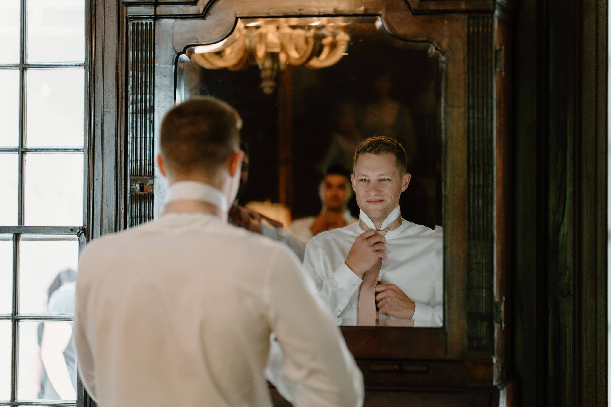 image over the groom's shoulder, as he looks in the mirror and smiles while straightening his tie