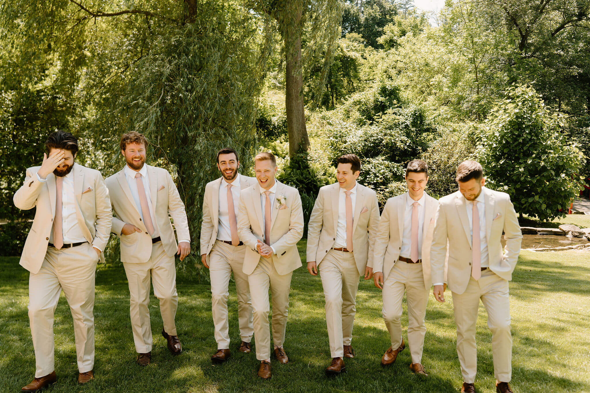 groom and groomsmen, all wearing tan suits with pink ties, laughing and walking toward the camera in a lush green yard
