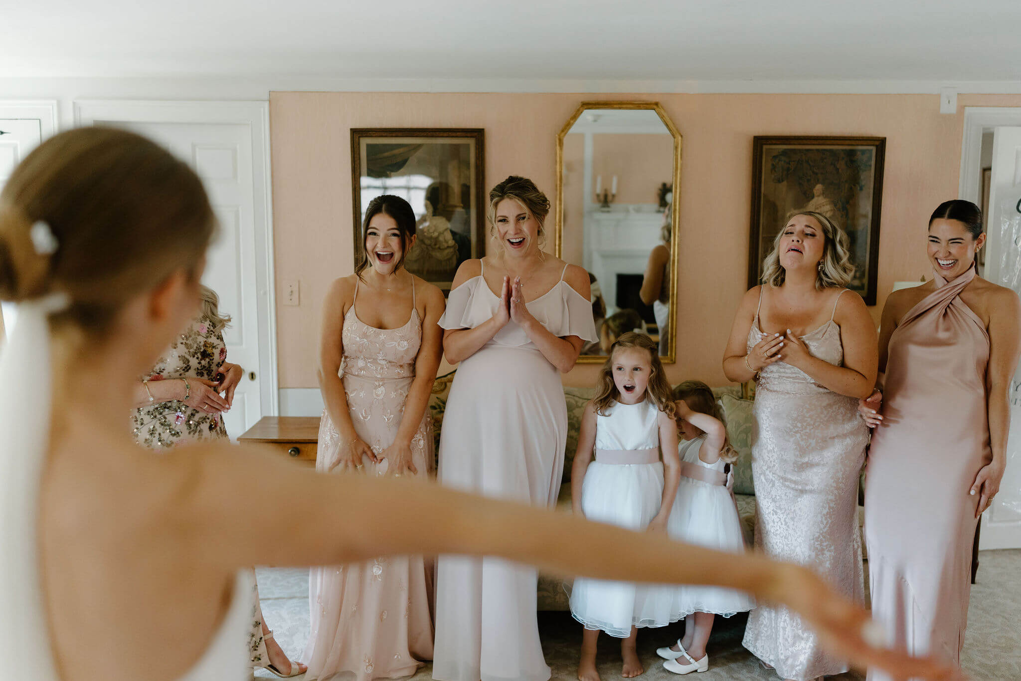 Bride with her arms wide open having a first look with her bridesmaids, all in various pink dresses, all smiling and delighted to see her