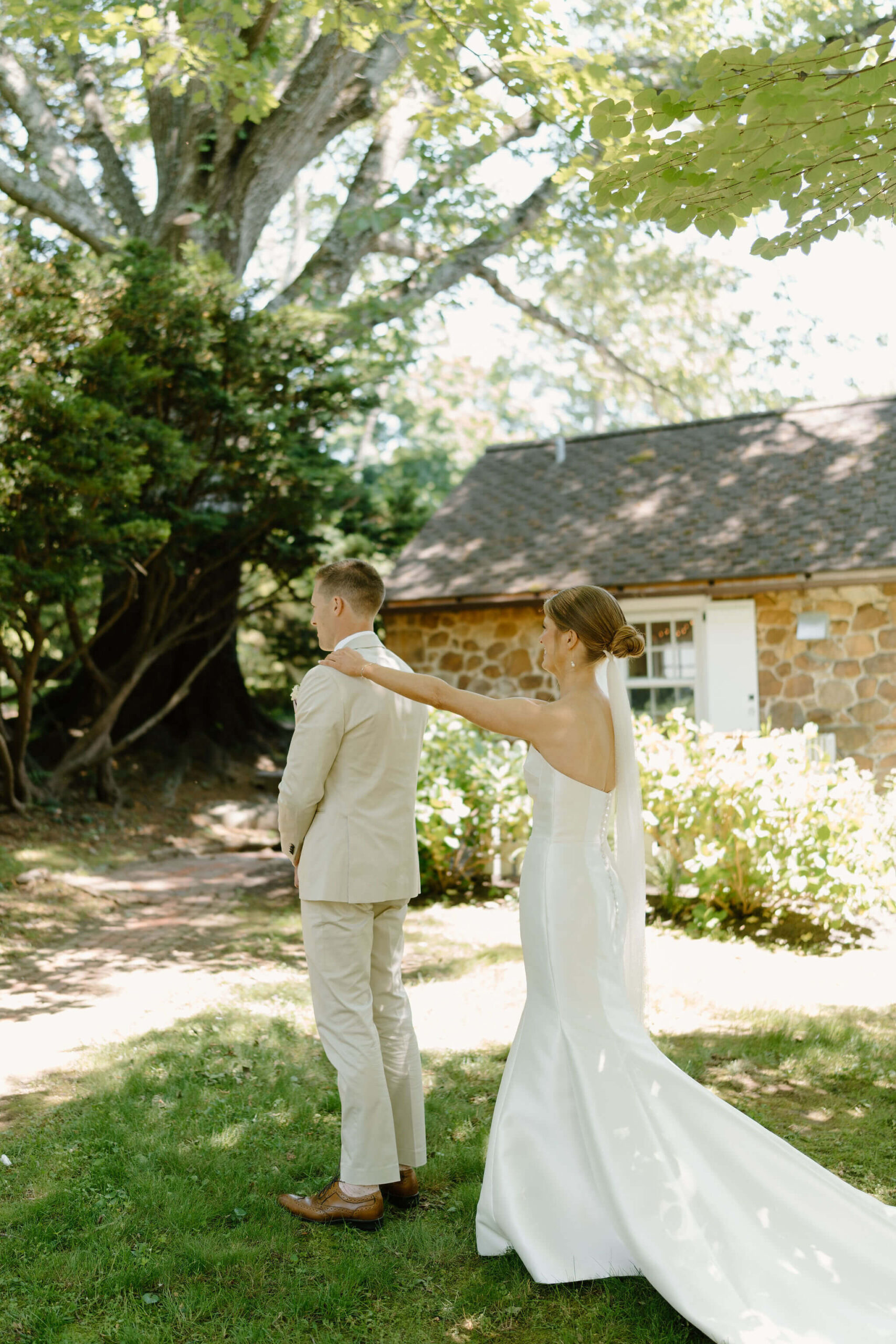 bride (white mermaid wedding dress) tapping groom (tan suit) on the shoulder for their first look, against a stone building with trees surrounding them