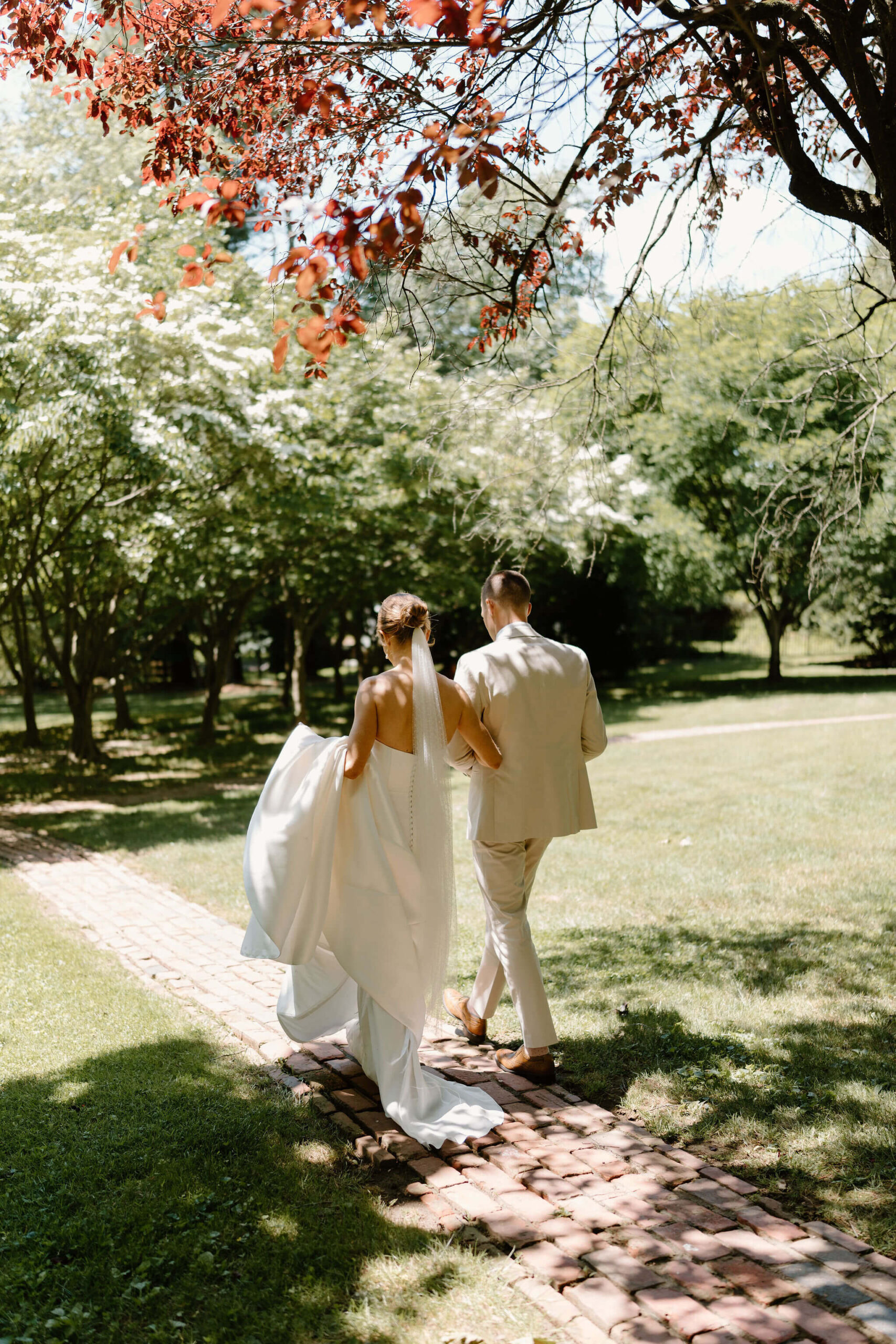bride and groom holding hands, bride holding her train, walking along a path toward trees