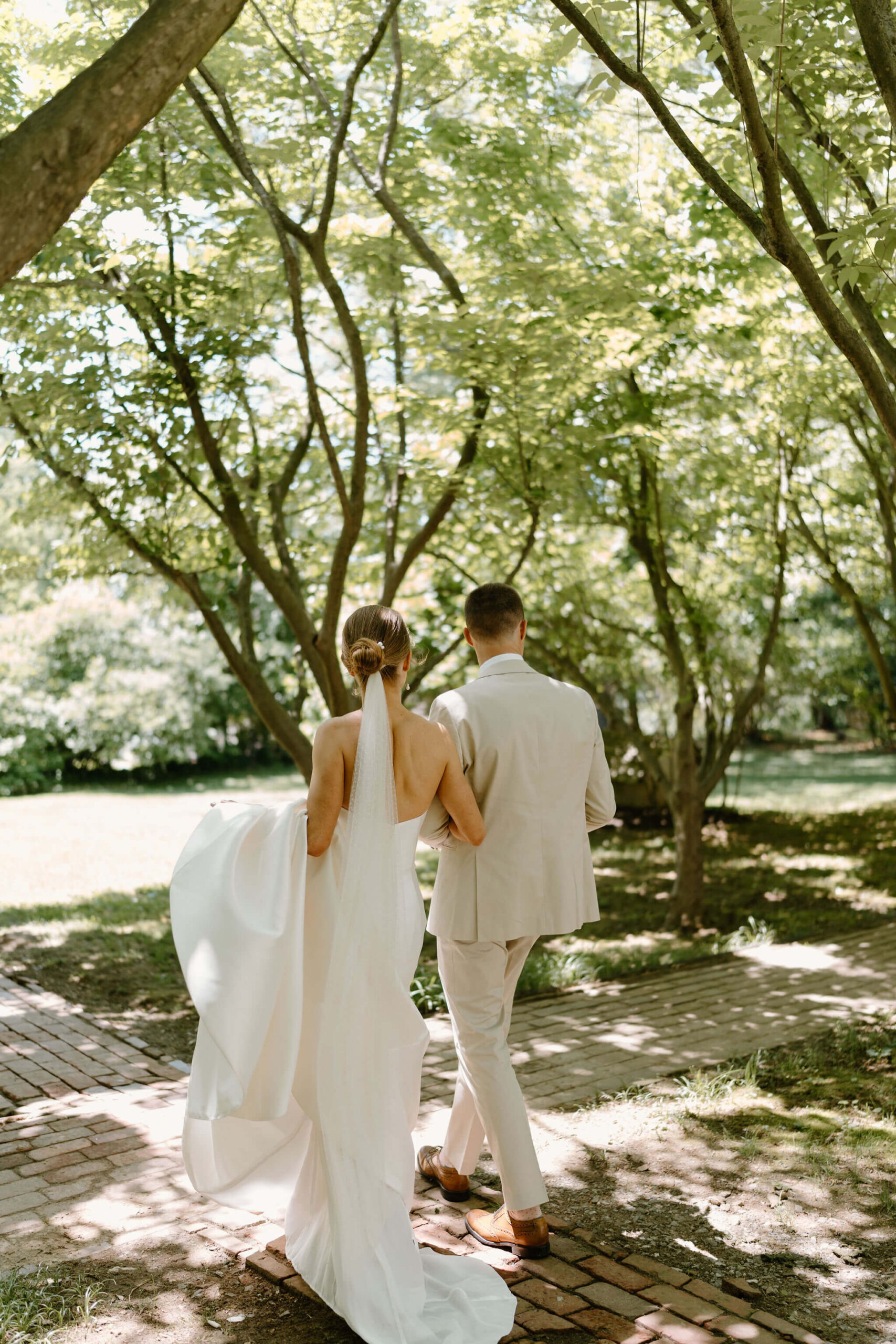 bride (holding the train of her wedding dress) walking with groom through a wooded path, away from camera