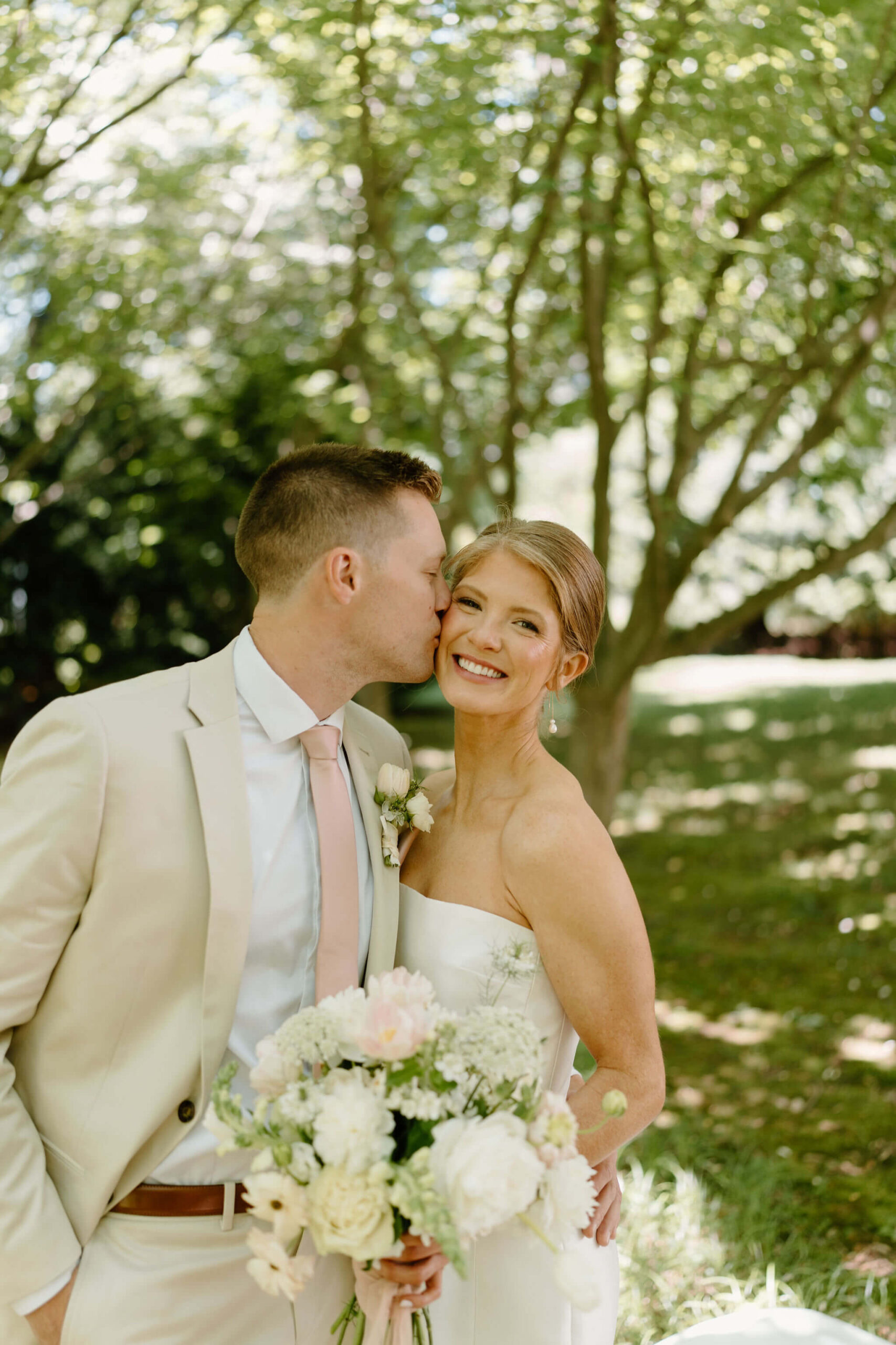 groom kissing bride on the cheek as she smiles toward the camera, holding her bouquet