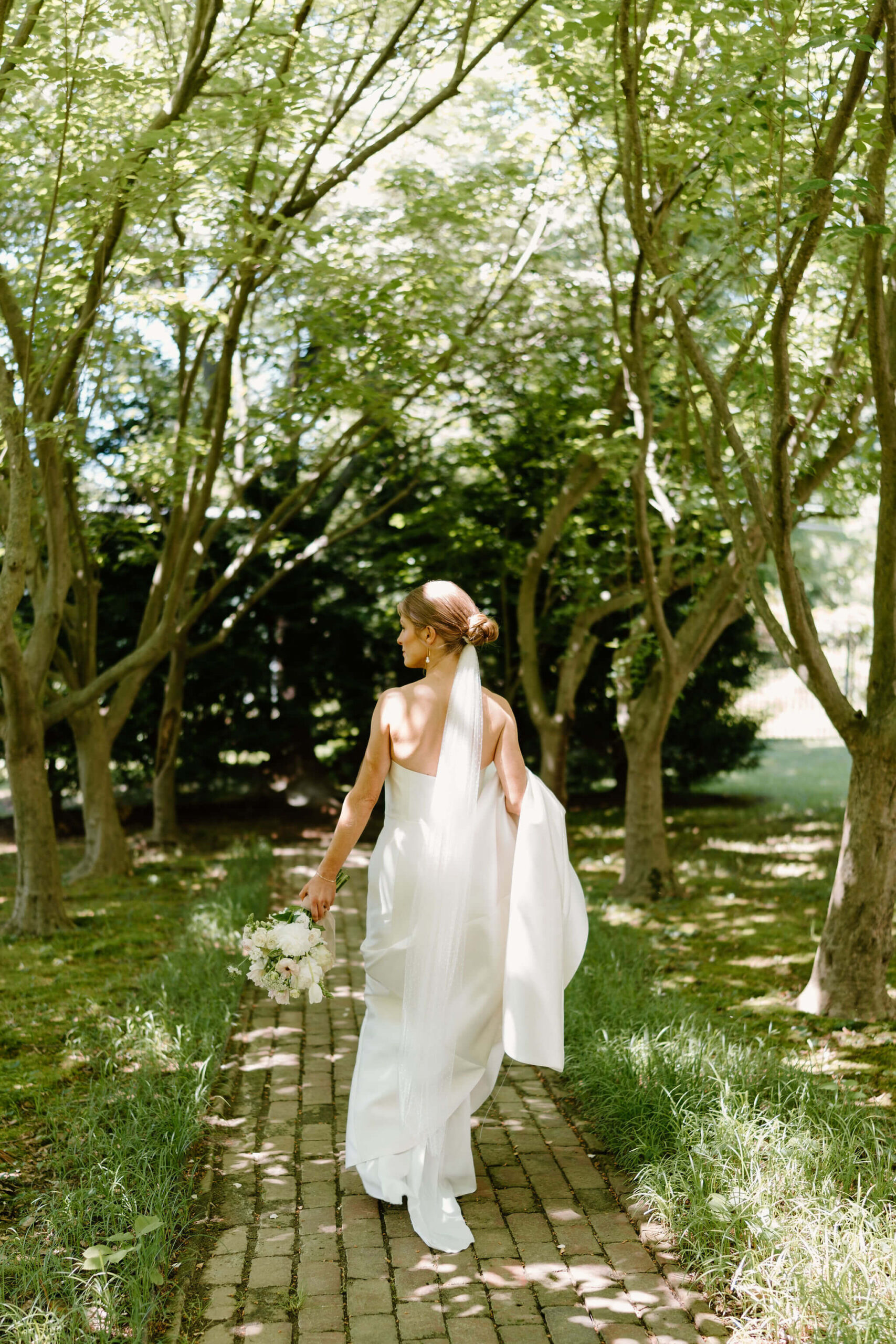 bride walking away from camera down a brick path underneath a canopy of trees, holding the train of her wedding gown with her bouquet down at her side