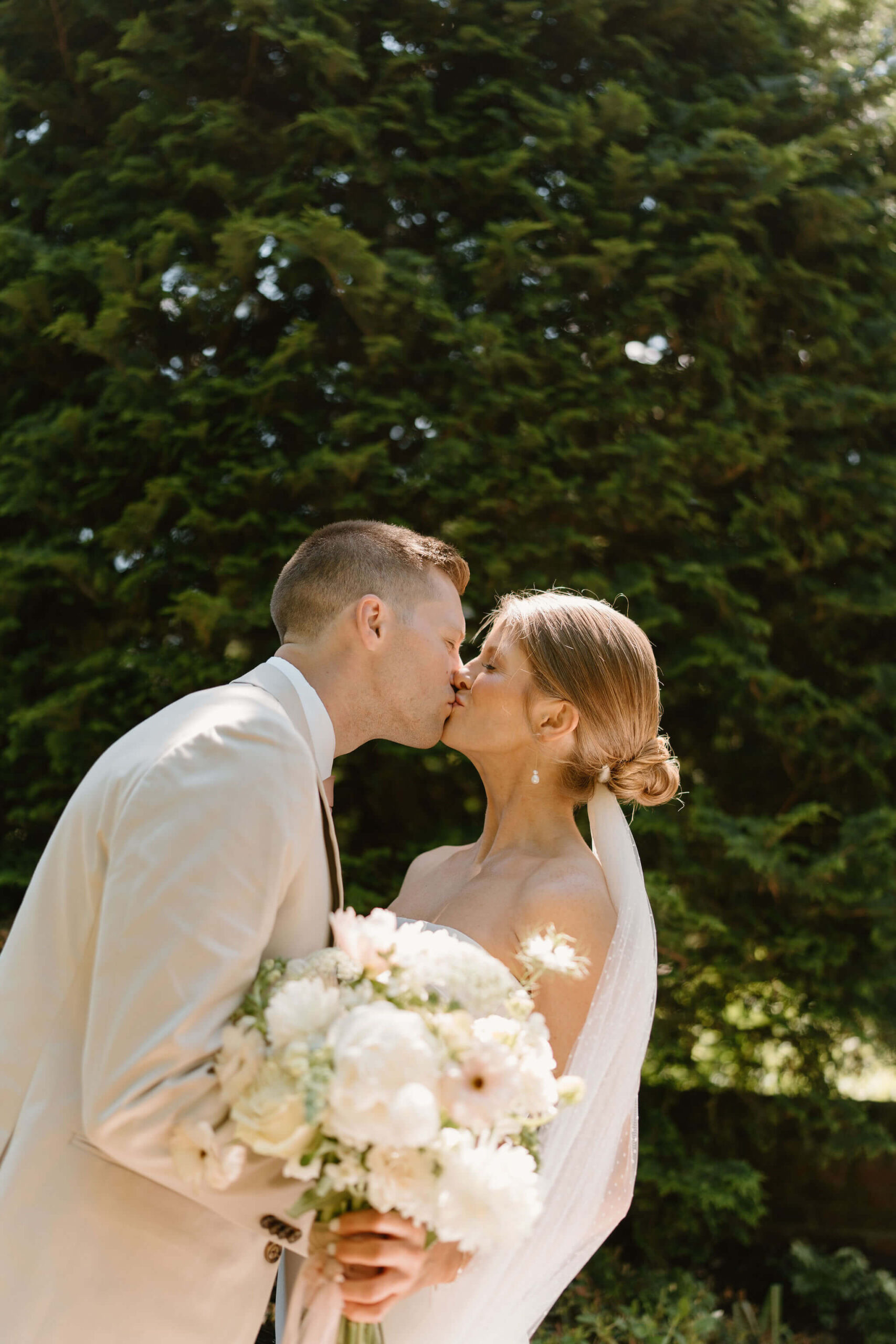 bride and groom kissing with her bouquet out in front of her, with an evergreen behind her
