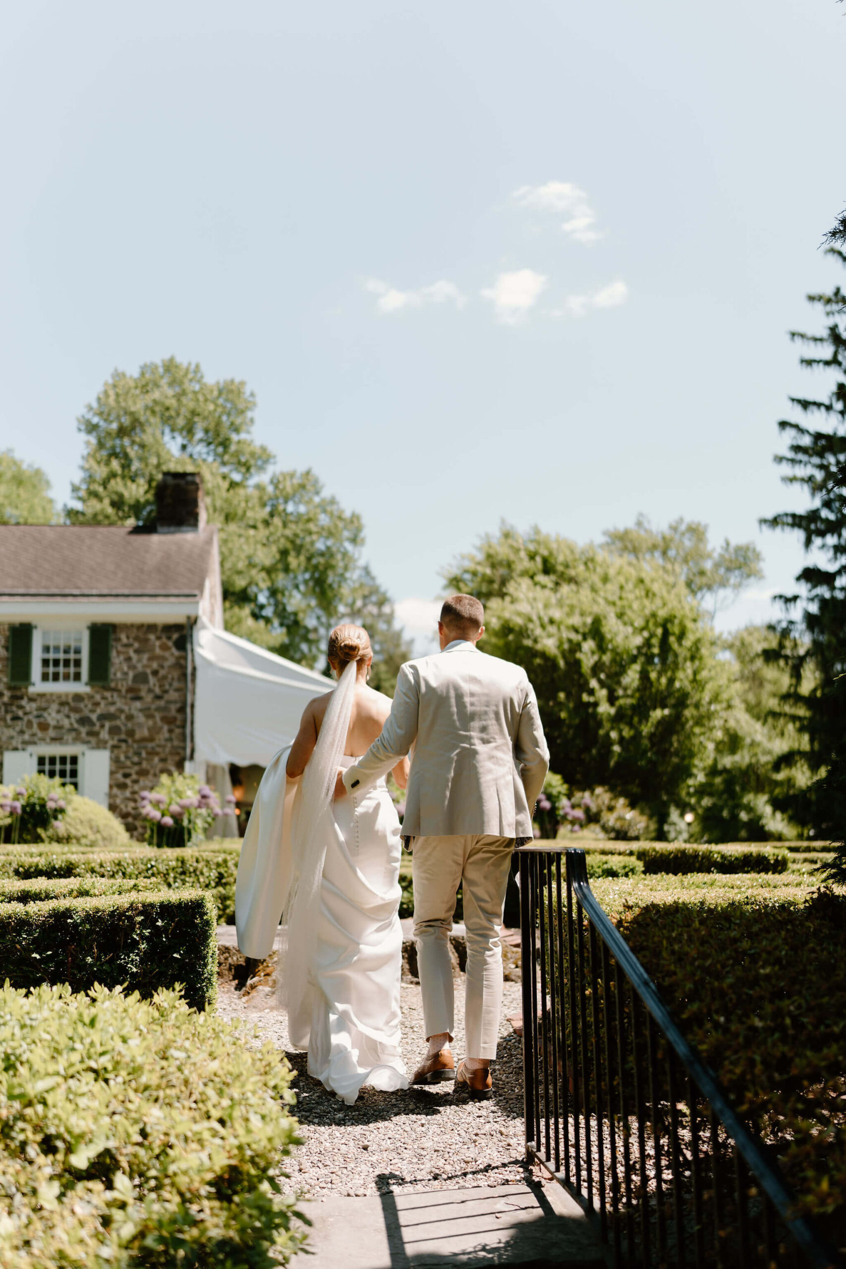 groom in tan suit with his hand on his wife's back as they walk through the hedge maze toward the stone house