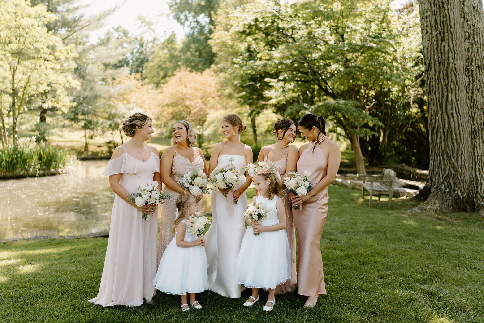 portraits of bride with her bridesmaids and flower girls, all wearing mismatched pink bridesmaids dresses, looking at each other and smiling against a lush green background