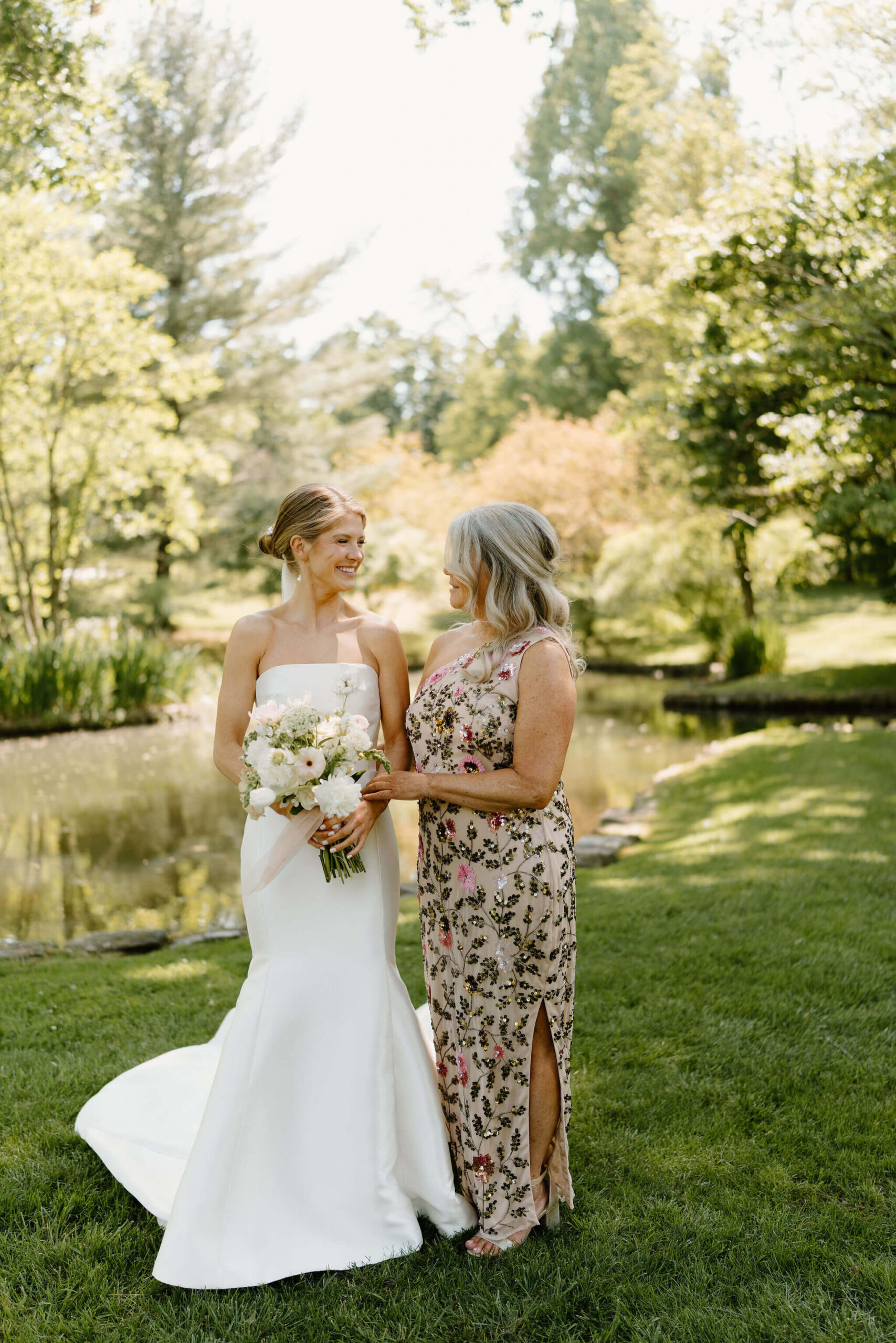 bride (white mermaid wedding dress, holding bouquet) smiling at her mother (pink dress with green floral sequined embroidery) for their spring wedding
