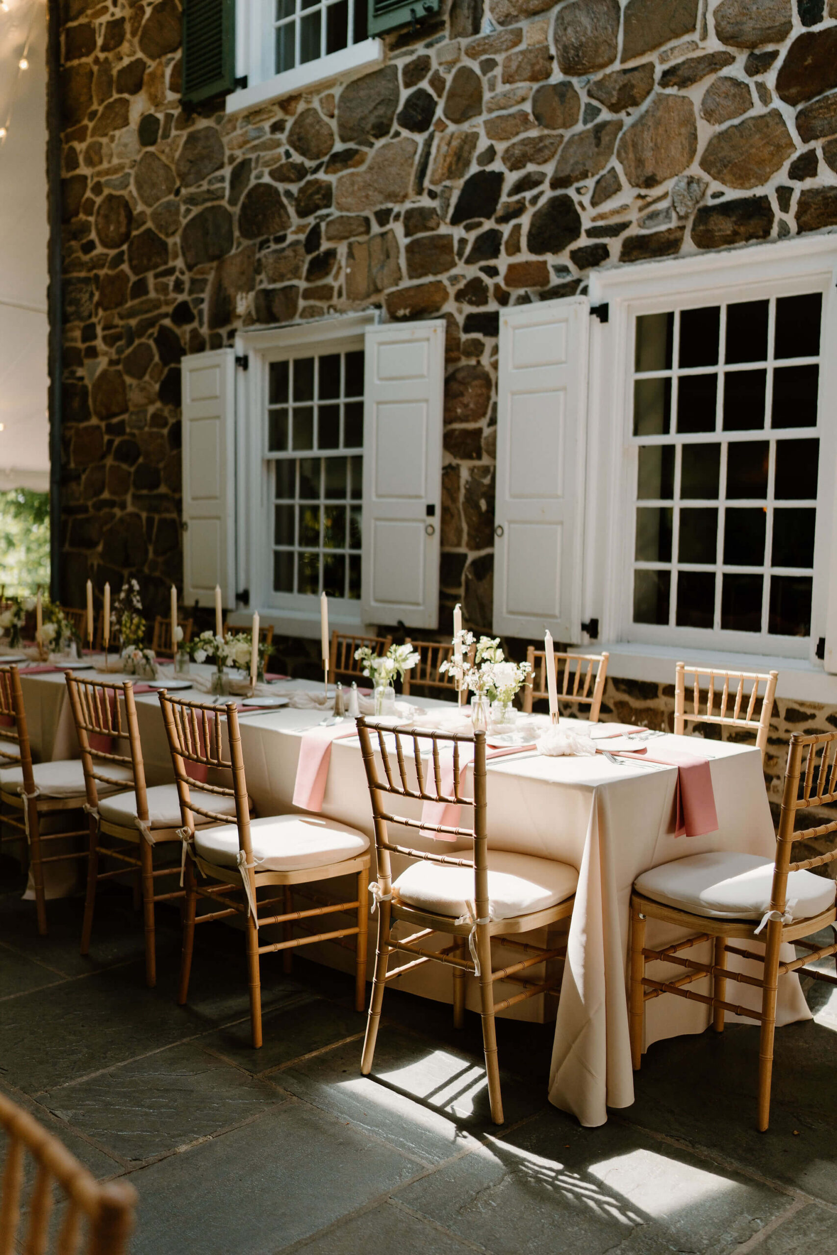 reception with wooden chairs in a sunny spot, against a stone building with white shutters. There are tall candlesticks and bud vases with small white flowers as centerpieces, with pink napkins at each place setting