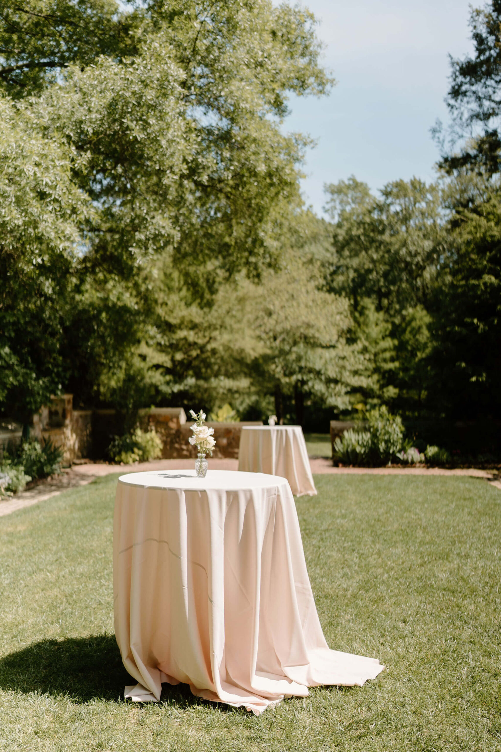 tow cocktail tables with white linens and a small floral centerpiece, in a large grassy courtyard in full sun