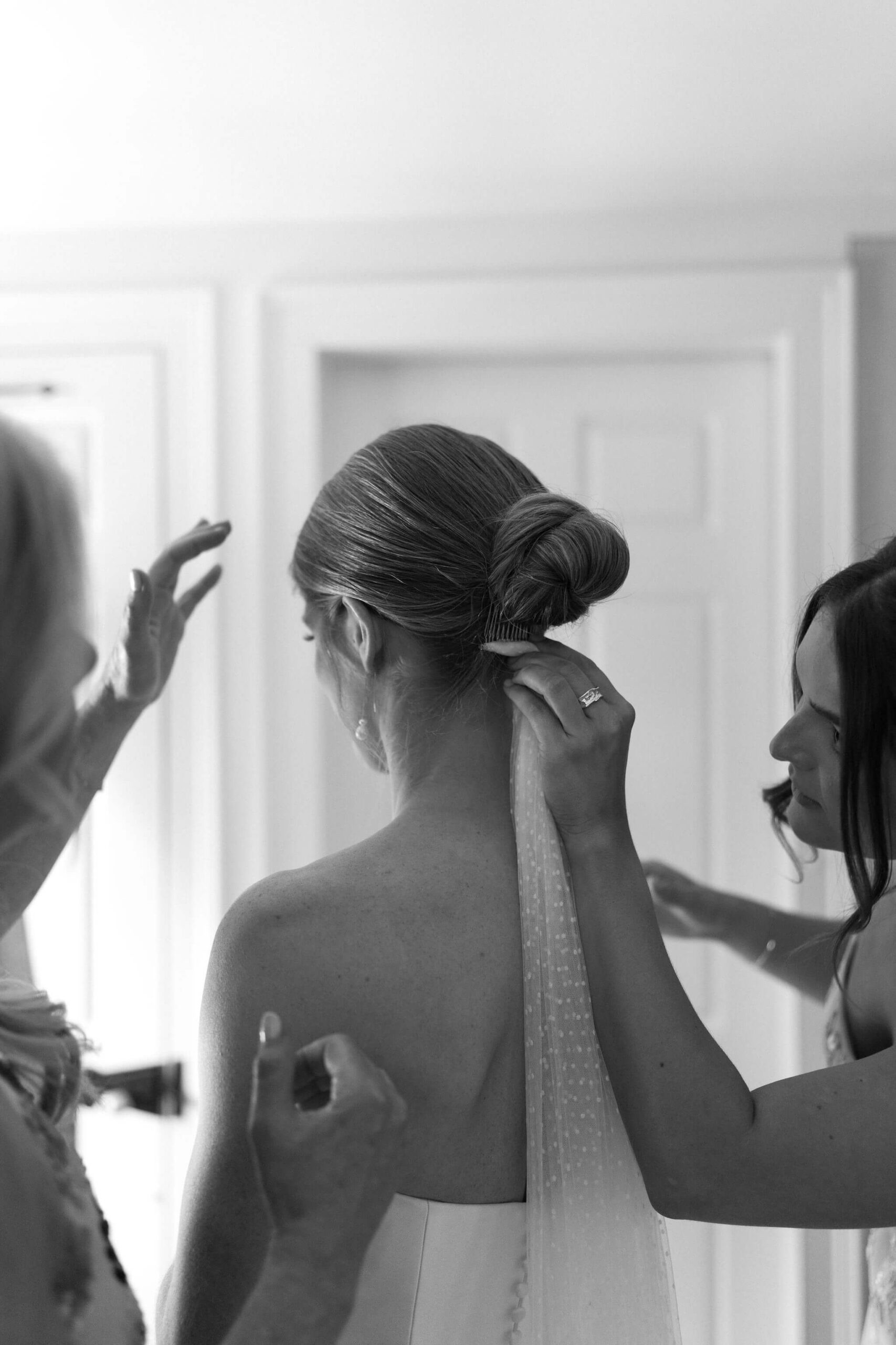 black and white image of the sister of the bride placing the bride's veil underneath her bun, while the mother of the bride reaches to help