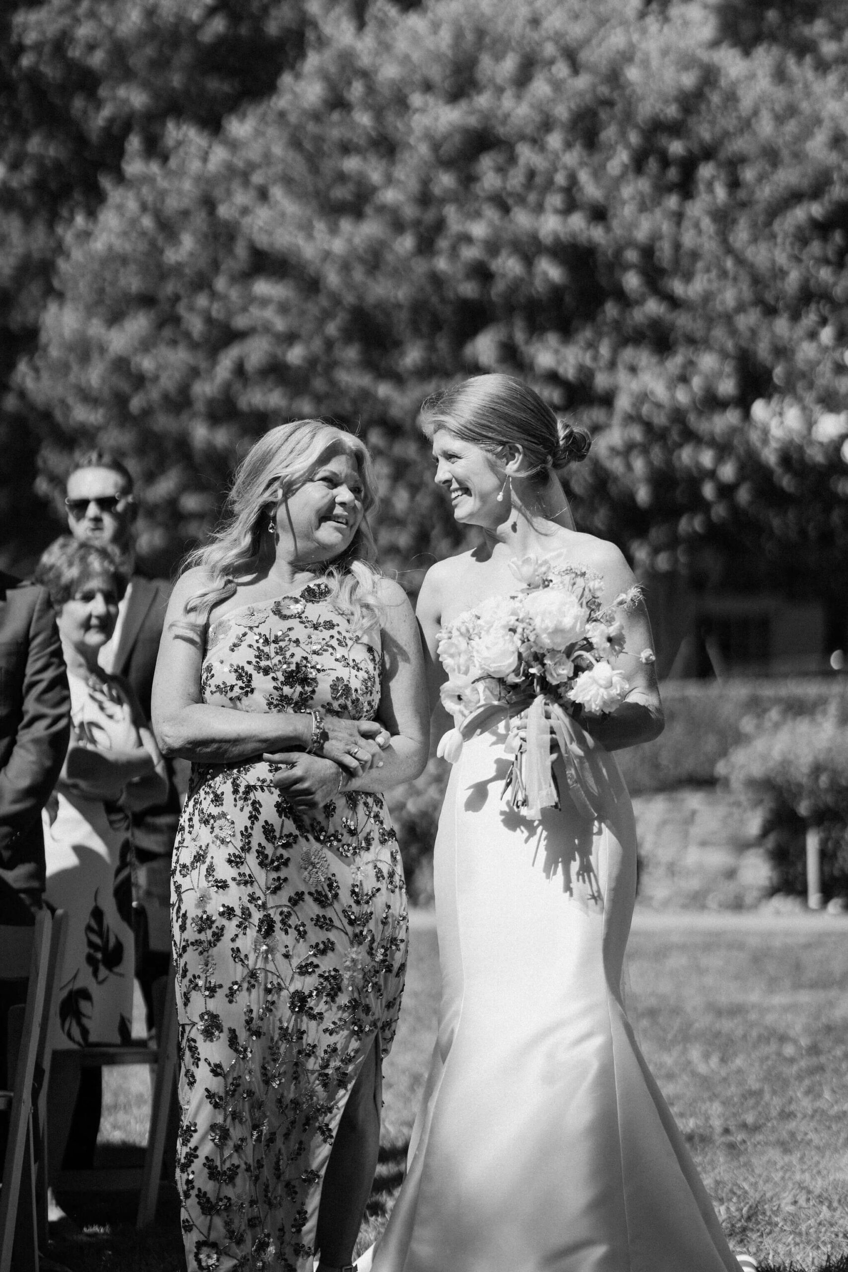bride and her mother smiling at each other as they walk down the aisle to her wedding ceremony