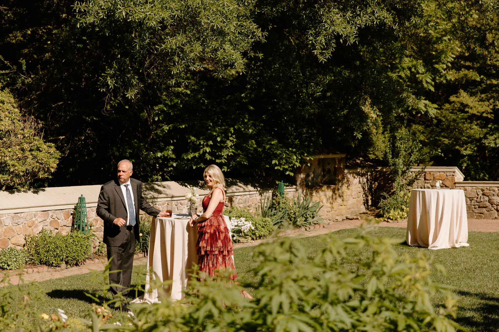 two guests standing at a cocktail table in a wide courtyard, smiling toward the cocktail hour area