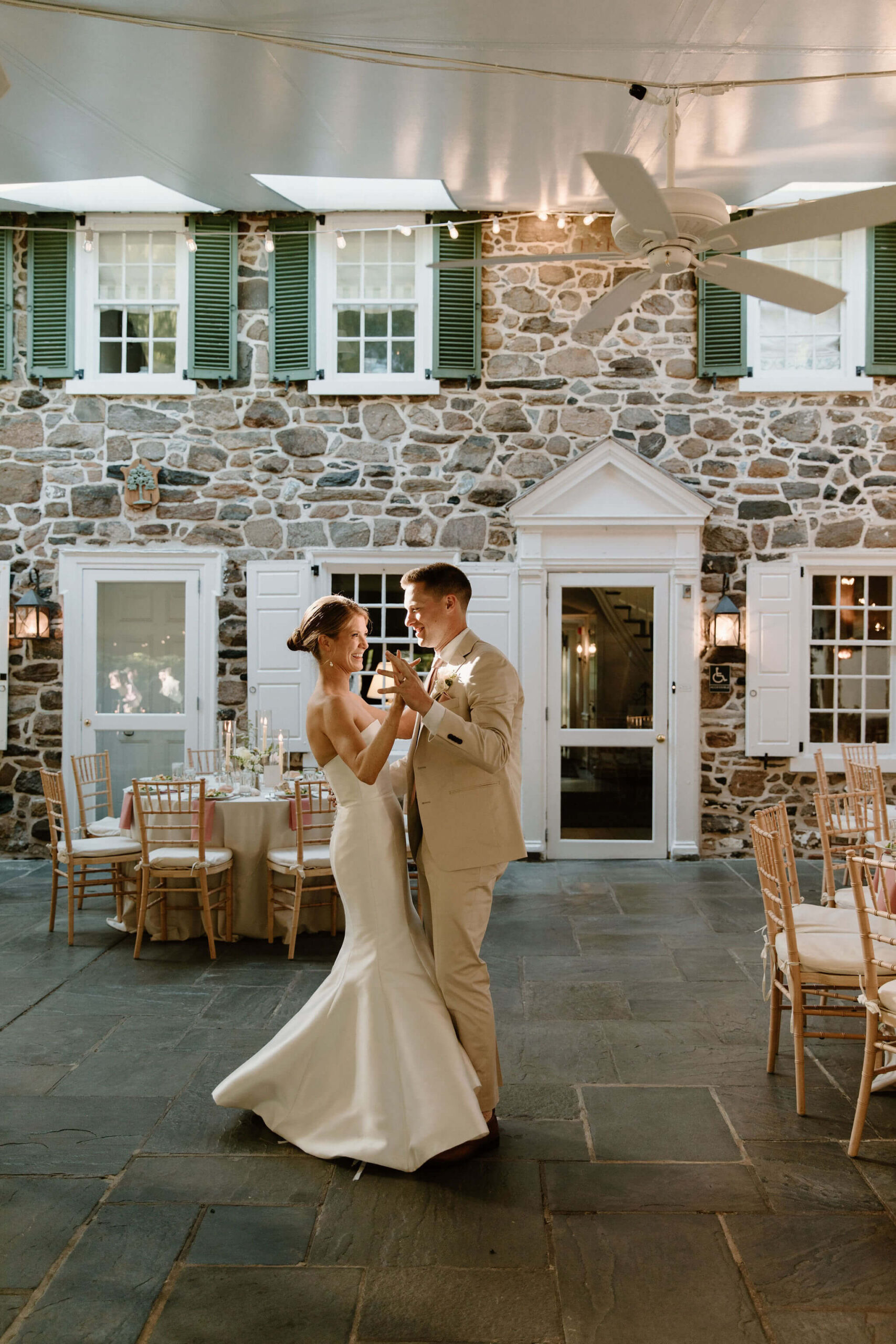 bride and groom practicing their first dance in their reception space, before any guests arrive, on a stone patio in front of a stone buildling