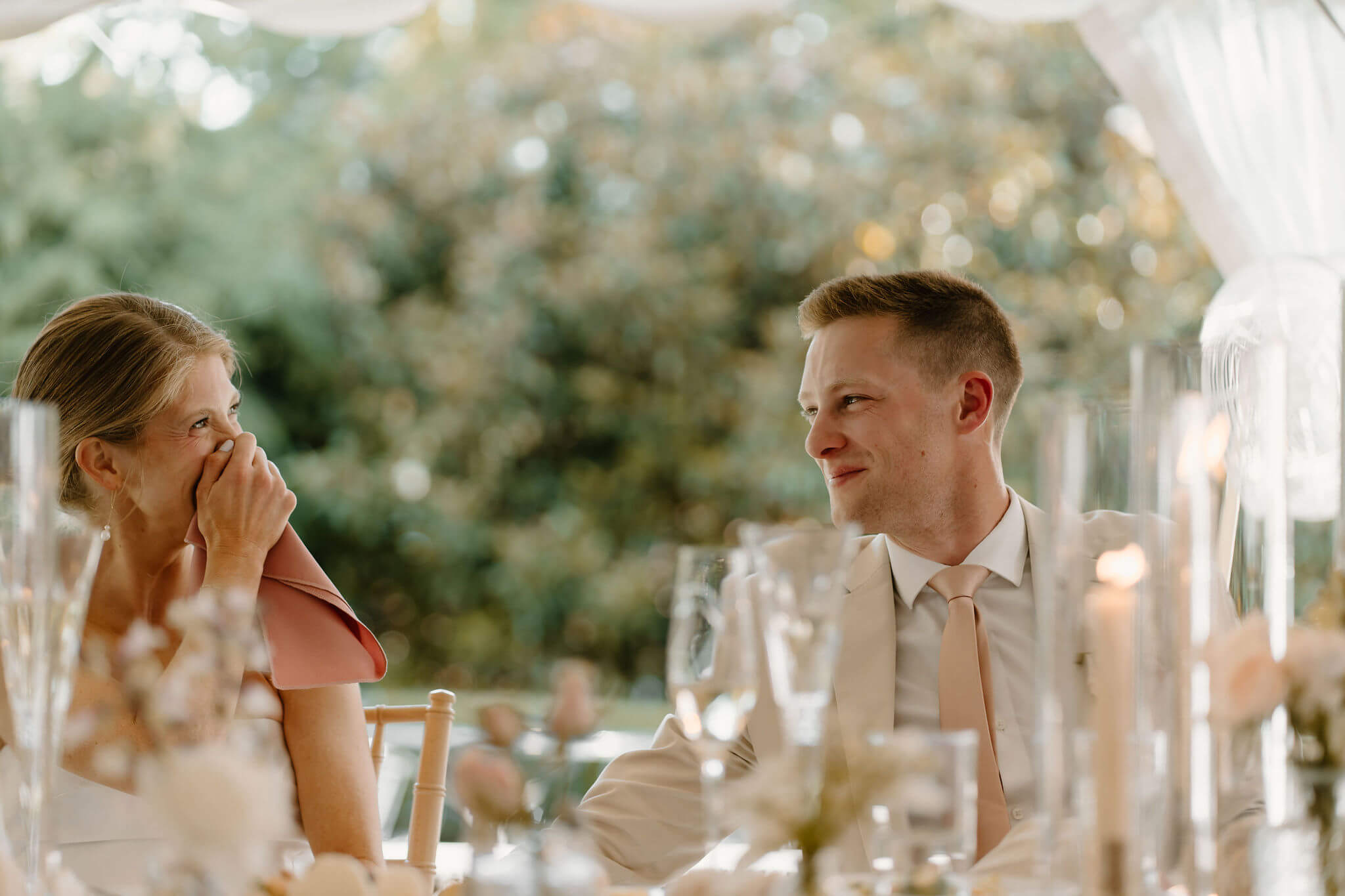 groom smiling at his bride as she covers her tearful, smiling face with a napkin during speeches